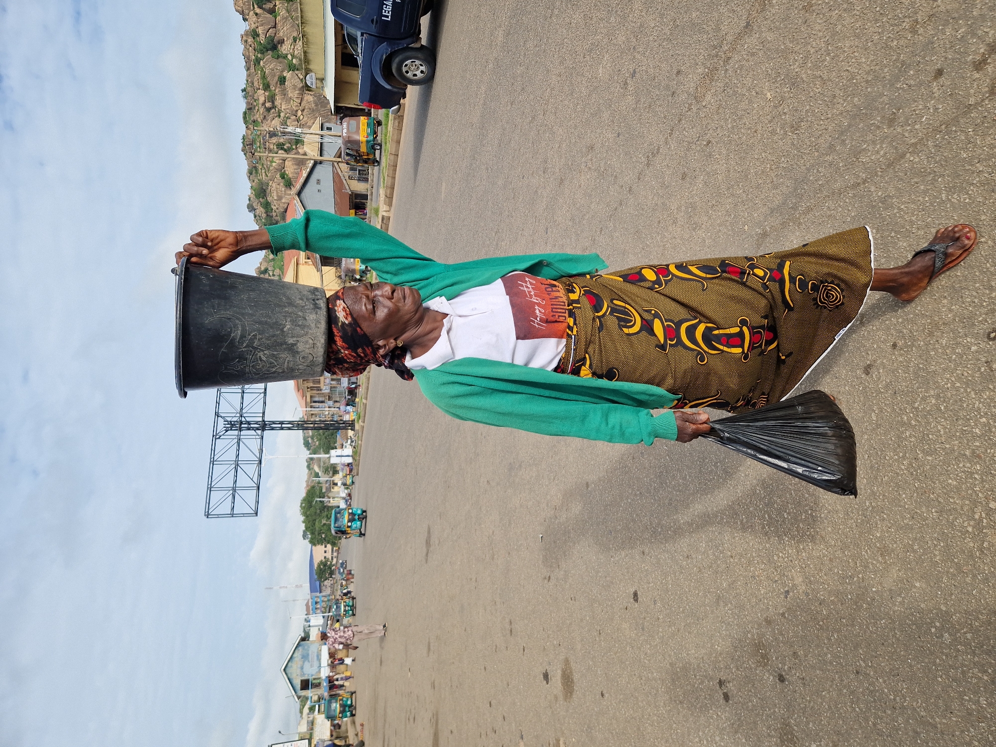 In the city of Kabong, Nigeria, a woman is walking on a paved road, carrying a large black bucket on her head. She is wearing a green cardigan over a white polo shirt with a graphic that reads "Happy Birthday" and "Squn". A patterned wrap skirt adorns her lower body. In her left hand, she carries a black plastic bag. The road is lined with buildings and shops on the left side, and rocky hills in the background. Several motorized tricycles, commonly known as 'Keke Napeps', are visible on the road, along with a dark blue pickup truck. The sky is overcast with a few clouds. The woman appears to be mid-stride, looking to her right.