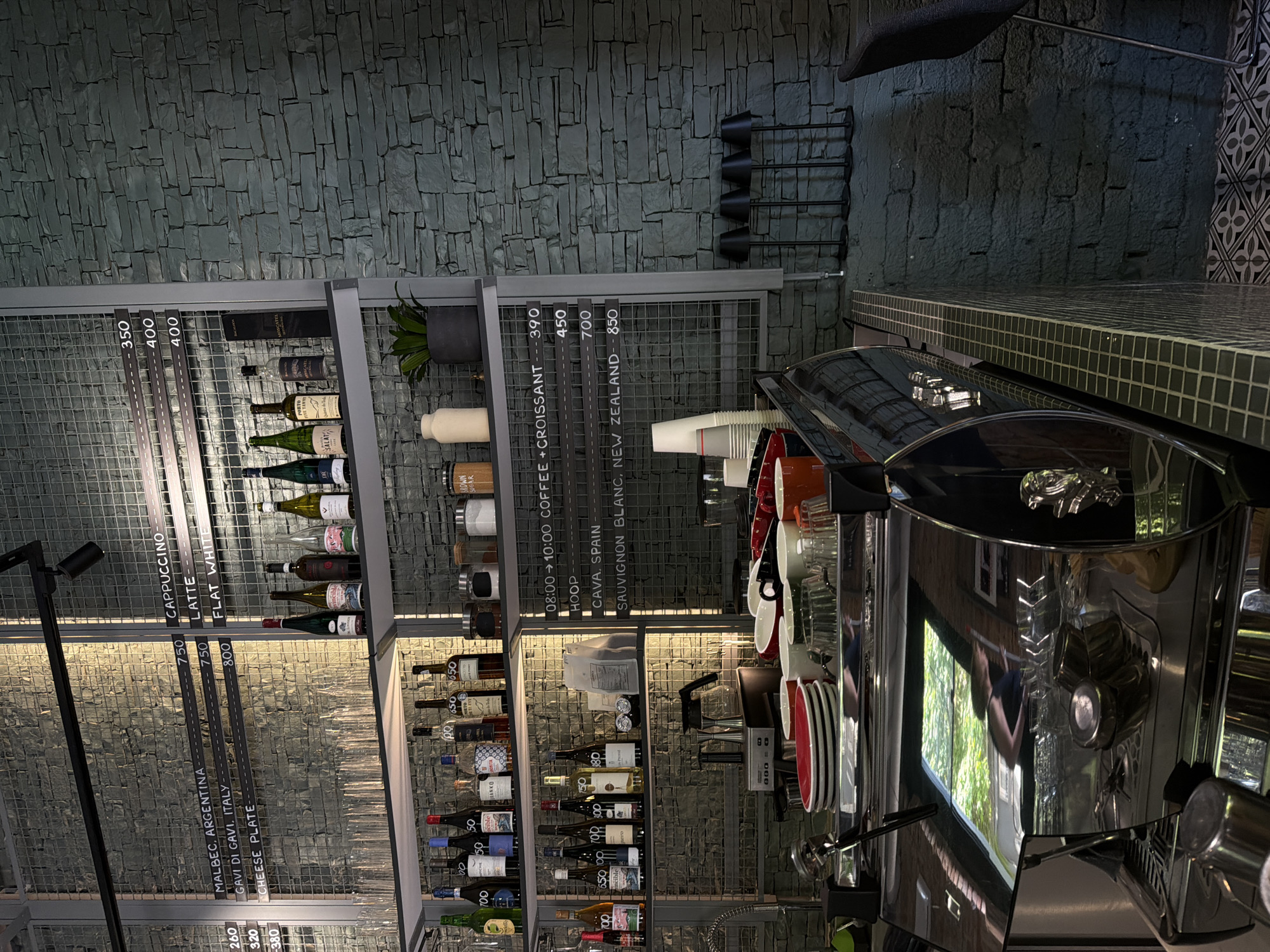 Here is a forensic description of the image:
The image shows the interior of a commercial establishment, possibly a cafe or bar. A modern, chrome espresso machine dominates the right side of the frame, reflecting the interior environment in its polished surface. Stacked beside the machine are cups and plates.
Behind the espresso machine, a metal wire shelving unit displays a variety of items. The shelves are stocked with bottles of wine, jars of coffee ingredients, and other culinary items. Notably, there is also a small plant in a gray pot.
Signage indicating a menu and pricing is affixed to the front of the shelves. Items listed include "CAPPUCCINO," "LATTE," "FLAT WHITE," "MALBEC, ARGENTINA," "GAVI DI GAVI, ITALY," "CHEESE PLATE," "COFFEE + CROISSANT," "CAVA, SPAIN," and "SAUVIGNON BLANC, NEW ZEALAND," each associated with a price.
The wall behind the shelves has a textured, stone-like appearance in shades of gray.
It is difficult to ascertain the precise location within Belgrade, Serbia from this image alone. However, the modern aesthetic of the establishment suggests it might be located in a contemporary or renovated area of the city.