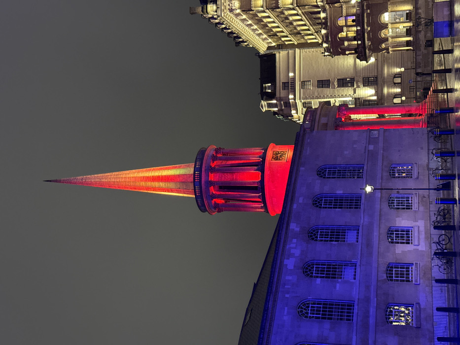 A tall, narrow spire, lit with orange and red lights, points upwards against a dark grey sky. It sits atop a cylindrical, pillared structure, also brightly lit. Adjacent to this, the side of a stone building with arched windows is illuminated in blue. Above, classic architecture is visible, bathed in warm yellow light. The perspective is tilted, giving an unusual angle to the urban scene.