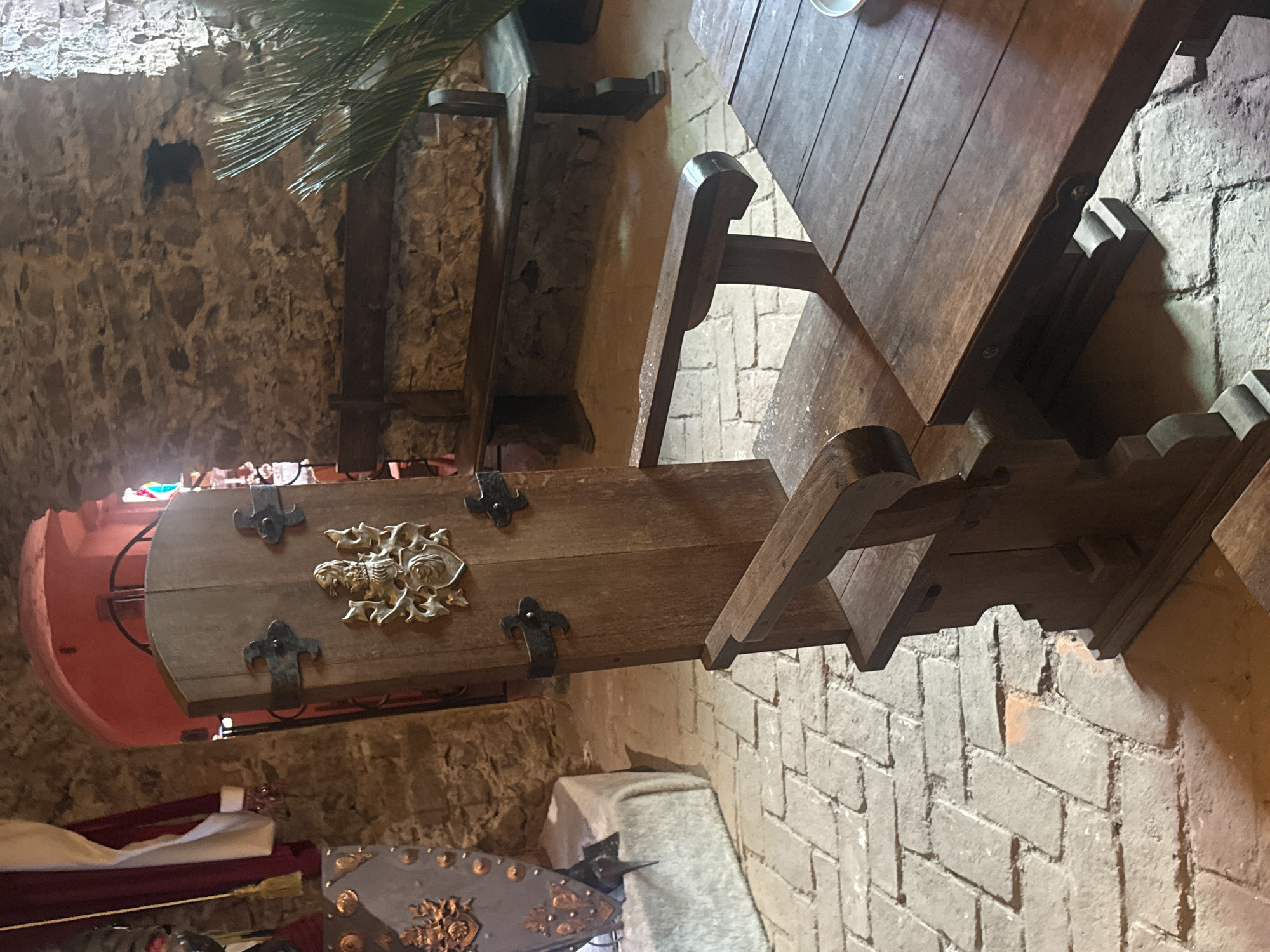 The photograph shows an interior setting, likely a room within Saint Miklós Castle in Chynadiiovo, Ukraine. The walls appear to be constructed of rough-hewn stone. A wooden table with carved legs and a matching bench are visible. The bench is adorned with metal fittings and a decorative emblem. A portion of a red-painted object can be seen, potentially a decorative element or part of a larger display. There are other decorative items like a shield with ornaments visible in the lower part of the image. The setting suggests a historical or medieval theme.