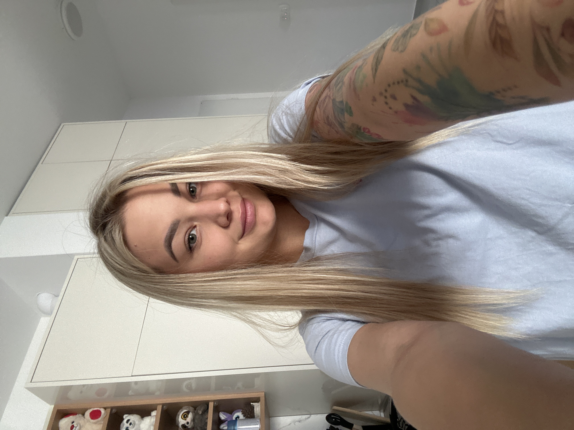 A young adult female is captured in a selfie taken indoors in Budva, Montenegro. The image is rotated 90 degrees clockwise, showing her head to the left and her body extending towards the right. She has long, straight, blonde hair that cascades around her shoulders. She looks directly at the camera with a slight, gentle smile, showcasing her green/hazel eyes and full, light pink lips. She is wearing a light blue or grey short-sleeved t-shirt. Her right arm, visible on the right side of the frame, is adorned with a colorful sleeve tattoo featuring abstract and floral designs in pastel shades of green, blue, pink, purple, and brown.
The background consists of modern, light-colored, possibly white or off-white, modular cabinetry. Above the cabinets, a plain white ceiling with a single recessed light fixture is visible. Below the cabinetry, a light wooden shelf unit with multiple compartments holds various stuffed animals, including a white teddy bear with red details, a grey sloth, and other plush toys. A small section of what appears to be a light-colored, possibly marble-patterned countertop or surface, along with some dark objects, is visible at the bottom right. The scene is brightly lit, suggesting natural daylight. No visible text is present. The overall atmosphere appears casual and relaxed.