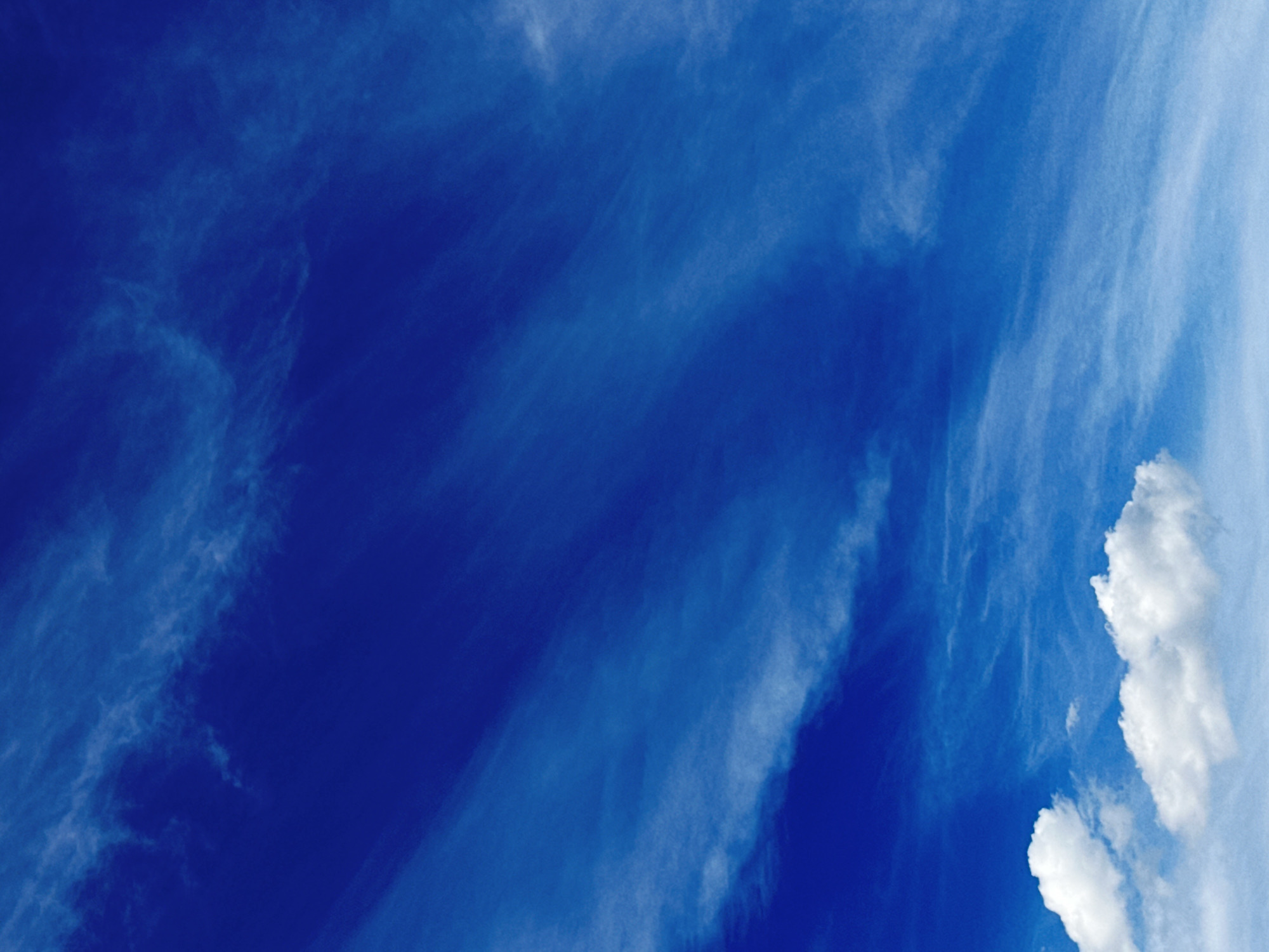 The image is a close-up, vertical, outdoor shot of a bright blue sky with scattered white clouds. The clouds are wispy and feathery, some forming thin streaks that stretch across the frame, giving the impression of wind. The overall tone is serene and airy, with the deep blue of the sky dominating the composition. No people, structures, or discernible ground features are visible.