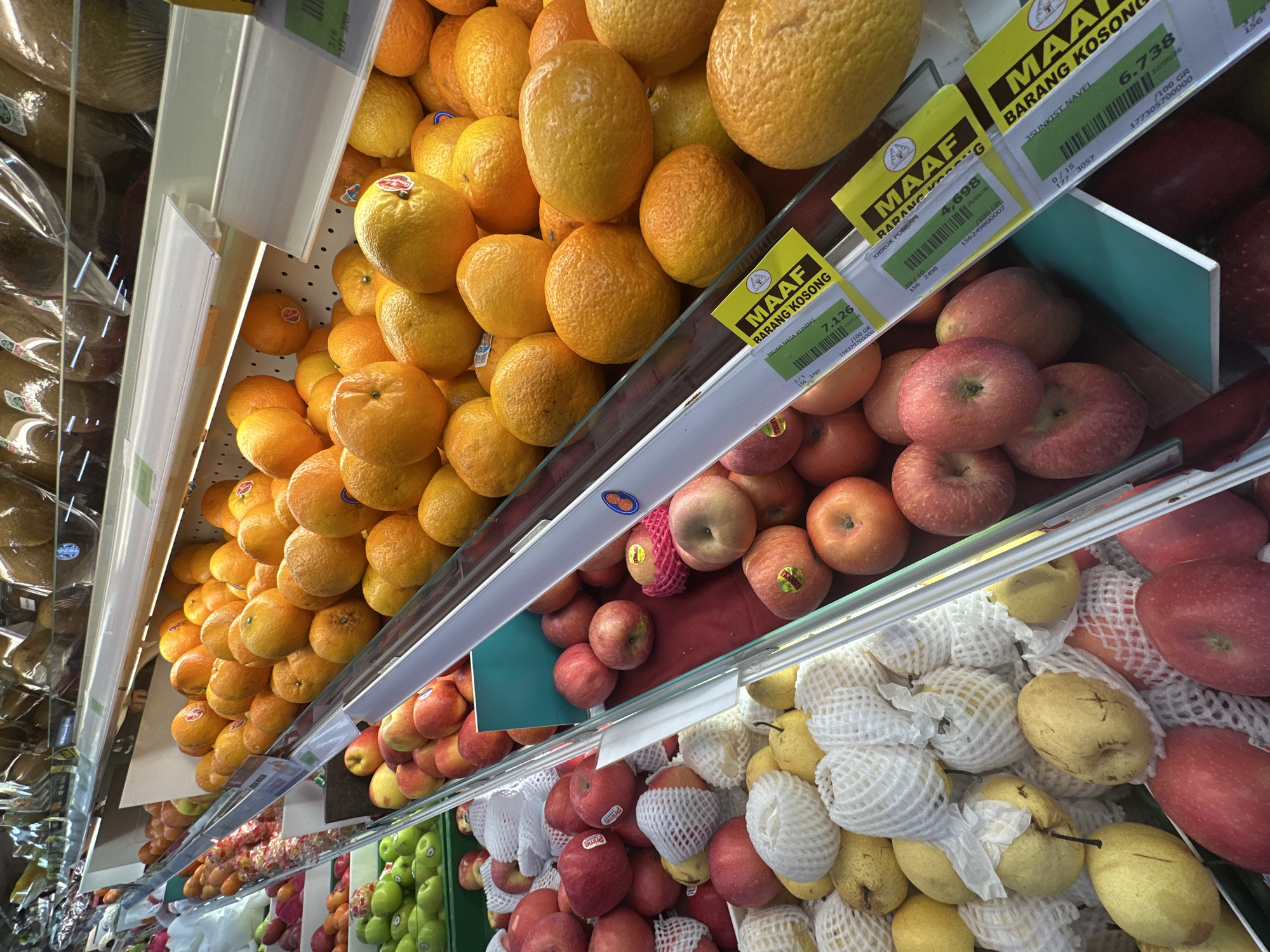 The image shows a produce aisle in a supermarket in North Kuta, Indonesia. The aisle is stocked with a variety of fruits displayed on shelves. In the foreground, red apples and yellow pears, some individually wrapped in protective netting, are visible. Behind them, a large quantity of oranges are piled high, with yellow price tags in front of them. Further back, other fruits like green apples are also displayed. The lighting is bright, typical of a supermarket environment. There are no people visible in the image.