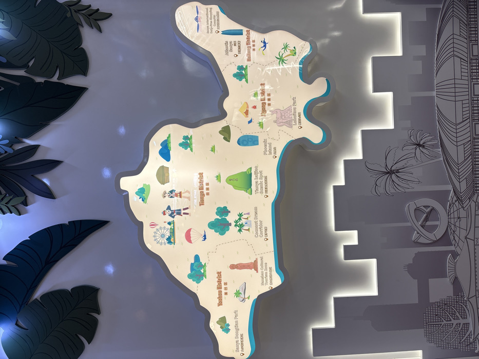 An indoor, illuminated wall display presents a stylized, three-dimensional map illustrating the districts of Sanya City, Hainan Province, China. The map, rendered in light tones with a prominent light blue lower border simulating a coastline, is centrally positioned and divided by dashed lines into distinct administrative areas, including Tianya District, Yazhou District, Jiyang District, and Haitang District.
Each district on the map features colorful, cartoon-style icons representing specific landmarks and attractions, accompanied by English and Chinese text labels with small locator pins. Tianya District, located towards the upper-center of the map, is depicted with a Ferris wheel, a parachutist descending, two stylized figures in local attire, and a traditional hut structure. Yazhou District, to the left of Tianya District, showcases icons for Sanya Dongtian Park, Nanshan Cultural Tourism Zone (a tall pagoda-like structure), Coconut Dream Corridor (palm trees), and Tianya Haijiao Scenic Spot (green rock formations). Jiyang District, situated below Tianya District, includes icons for Phoenix Island (a blue skyscraper-like building), a camping tent, and Luhuitou Park (a statue of a deer and figures). Haitang District, to the right, features icons for Sanya International Duty-Free Shopping Complex and Atlantis Sanya Resort (both represented by tall skyscrapers), and figures engaged in water activities.
Above the map, several layered, dark green, cutout shapes resembling tropical leaves are affixed to the wall, some illuminated by spotlights. Below the map, a multi-layered grey silhouette artwork depicts a modern cityscape, including numerous buildings, palm trees, and a large, stylized infinity symbol. This cityscape element is backlit along its upper edge, creating a glowing separation from the map above. The entire display is mounted on a uniform, light-colored wall, utilizing internal backlighting for the map and cityscape, along with external illumination for the foliage elements.