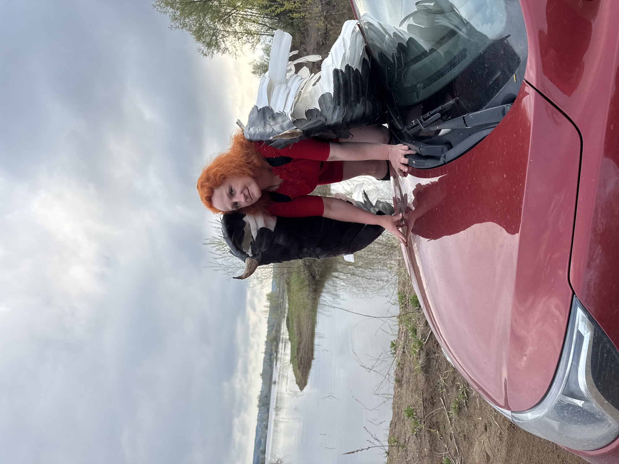 The image shows a woman with red hair wearing a red dress with wings attached, kneeling on the hood of a red car. The car is parked on a patch of dirt and grass near a body of water. The background includes trees and a cloudy sky. The location appears to be Noviy, Russia.