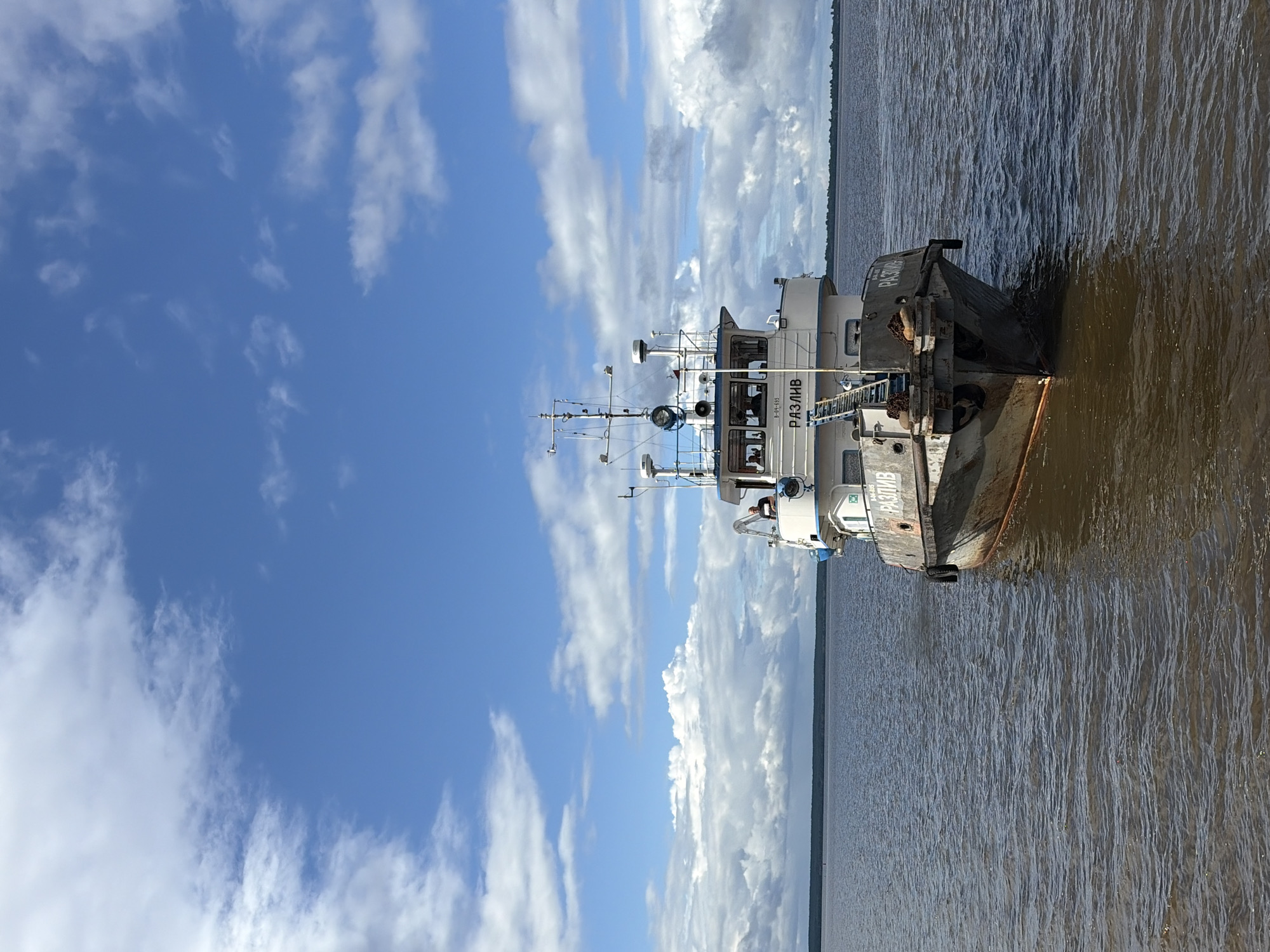 This is a vertical, eye-level shot of a ship on a wide, murky river. The sky above is a bright blue with scattered, fluffy white clouds. The river is a brown-grey color with small ripples on the surface. The ship is white with some dark grey accents and appears to be a working vessel. It has a raised cabin with windows and various antennas and equipment on top. A name is visible on the side of the ship, written in white Cyrillic letters: "РАЗЛИВ". There is a ladder extending from the ship to a darker, flat-topped barge that is partially submerged in the water. The barge also has some visible text, though it is less clear. The lighting suggests it is daytime, possibly late morning or early afternoon, with bright sunlight casting shadows. The overall scene is one of a calm day on a large body of water, with the ship and barge appearing to be either docked or navigating slowly. There are no people clearly visible.