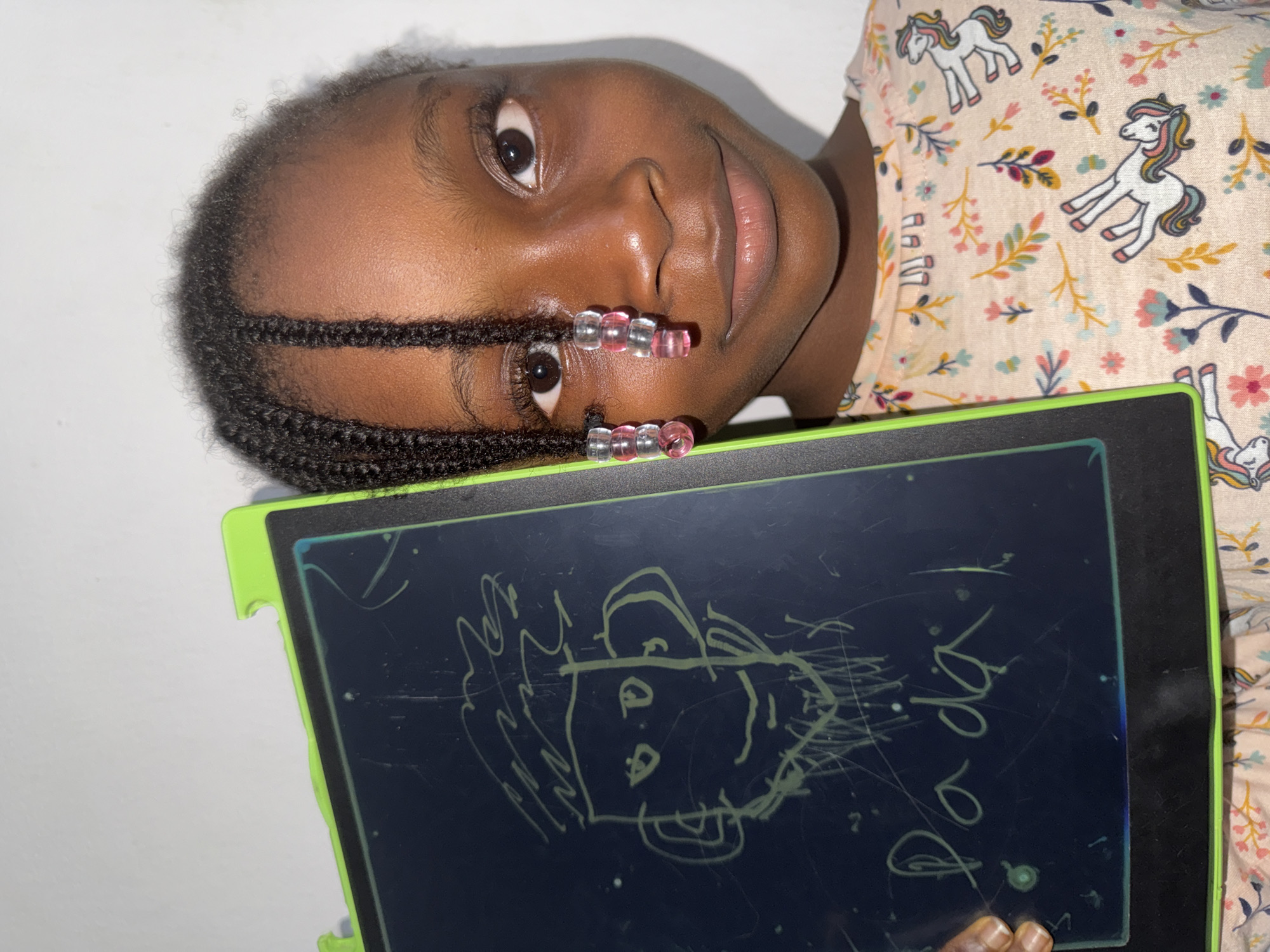 The image captures a young Black girl, likely in Ado, Nigeria, positioned at an angle as if reclining against a plain light-colored background. She looks directly at the camera with a calm, pleasant expression and a slight smile. Her dark braided hair is decorated with several translucent pink and clear beads, forming a decorative line along her scalp.
She is wearing a light-colored short-sleeved top adorned with a cheerful pattern of white unicorns featuring colorful manes and tails, interspersed with various small, brightly colored floral and leafy designs.
In her hands, the girl holds a dark-screened LCD writing tablet with a bright green frame. The tablet's surface displays light-green line drawings. A central drawing depicts a simple, childlike caricature of a face or figure, characterized by scribbled hair, distinct eyes, and a broad smile. To the right of this drawing, the words "da da" are written in a simple, child-like script. Other faint scribbles are also visible on the screen. The scene is brightly lit, suggesting an indoor environment.