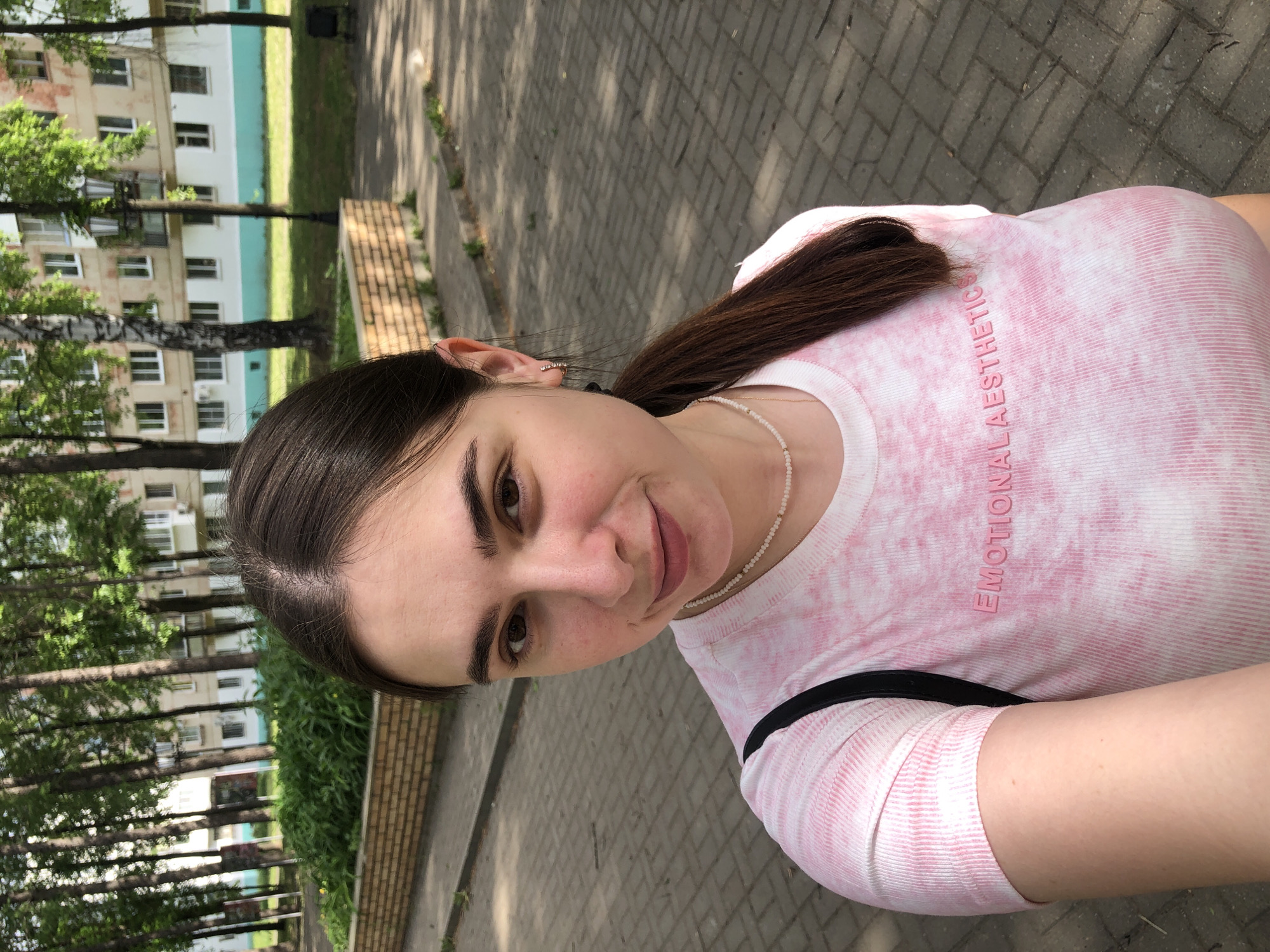 A woman is standing outside, likely in a park or urban area of Chaikovsky, Russia. She is wearing a pink and white tie-dye long-sleeved shirt with the words "EMOTIONAL AESTHETIC" on the front. She also has a white beaded necklace. Her dark hair is pulled back in a low ponytail. The background shows trees, a building with windows, and a brick sidewalk. The image seems to have been taken at an angle.