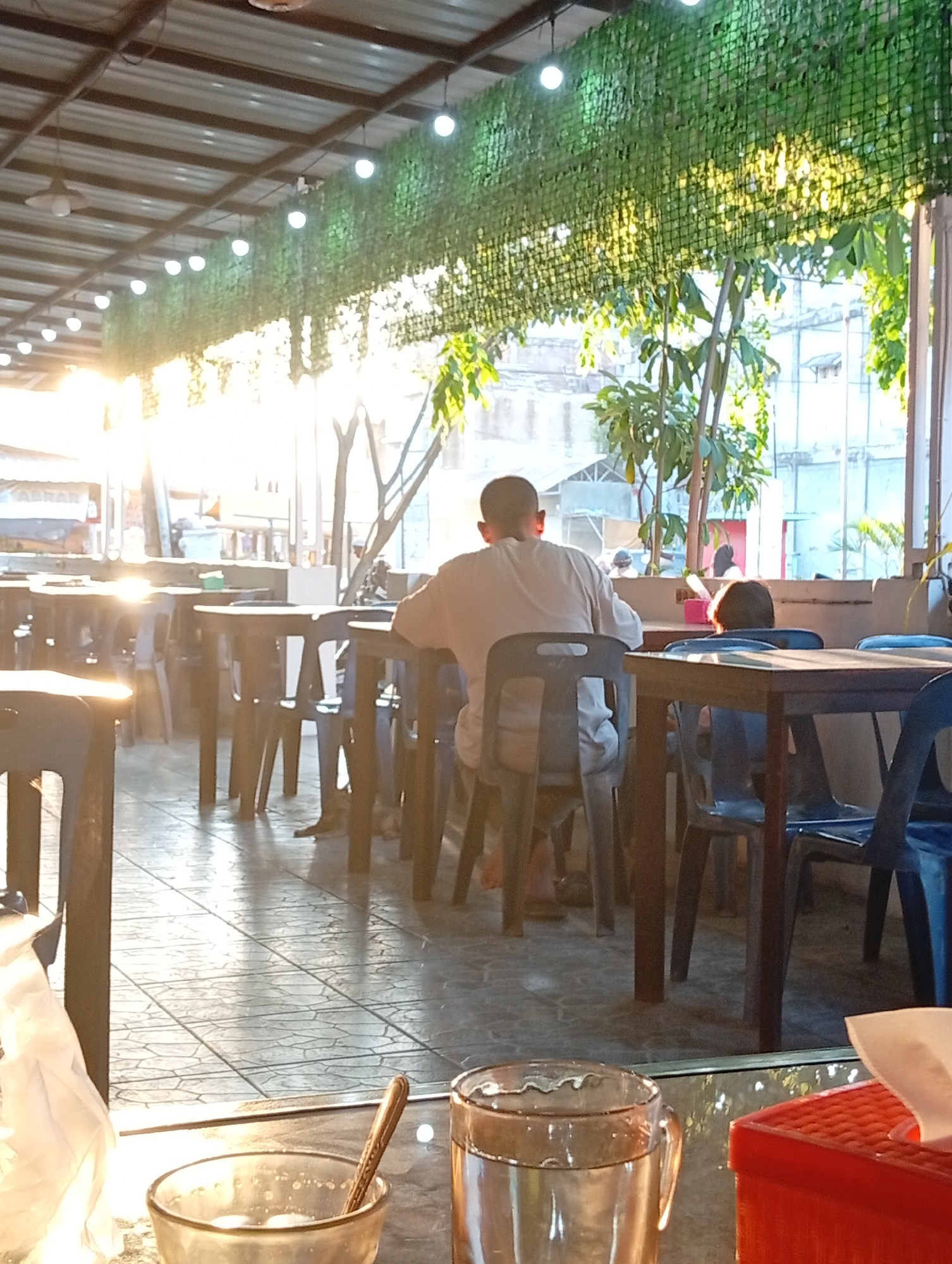 A man wearing a light-colored shirt is seated at a table in an outdoor restaurant. The man is facing away from the camera, looking towards a bright light source, possibly the sun. In front of him is a dark-colored chair. Several other tables and chairs are visible in the background, some occupied by other individuals, including a child. The restaurant appears to have a covered roof with lighting fixtures. The ground is paved with tiles. The scene is illuminated by warm, possibly sunset, lighting. A red tissue box and a glass of liquid are in the foreground, suggesting a dining or waiting area. The restaurant is located in Lhokseumawe, Indonesia.