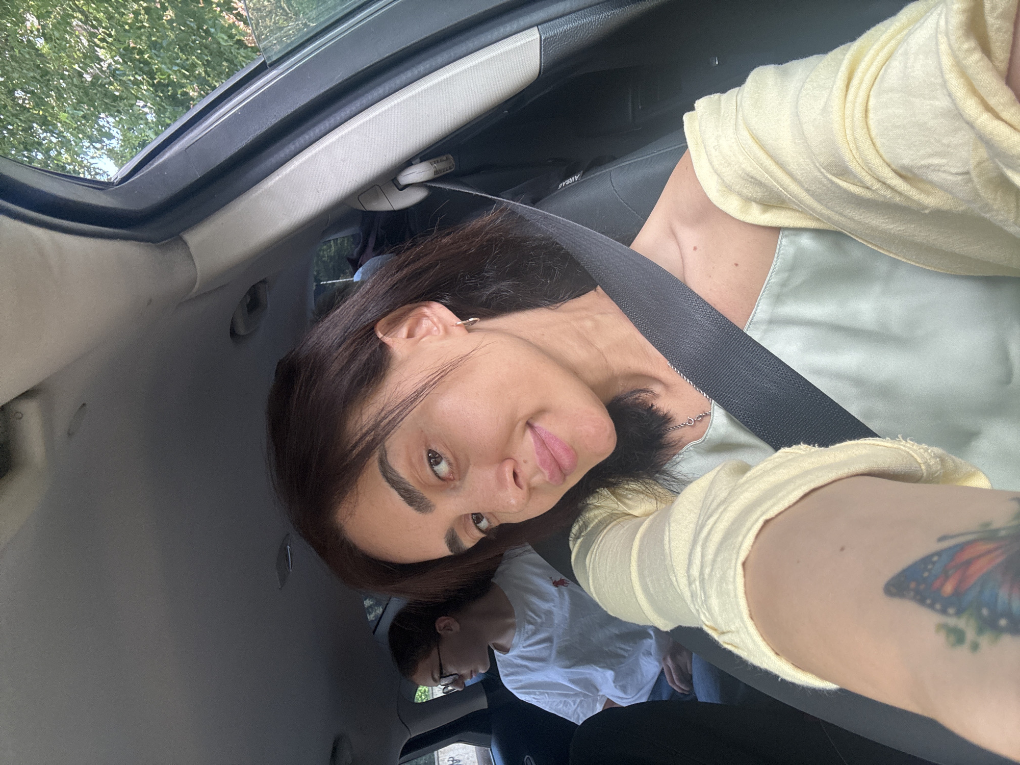 In a vehicle's passenger seat, a woman with dark hair and fair skin smiles at the camera, her left arm positioned to capture the selfie. She wears a light yellow off-the-shoulder top layered over a pale green camisole and a silver necklace. A black seatbelt is visible across her chest. On her left forearm, a tattoo of a butterfly with blue, orange, and black wings is partially visible. In the backseat, a young woman with glasses and dark hair pulled back wears a white t-shirt. The interior of the car is visible, with the ceiling and a portion of the right door frame. Outside the car, green trees and foliage suggest a natural, possibly suburban or rural, environment. The overall impression is a casual moment captured while traveling.