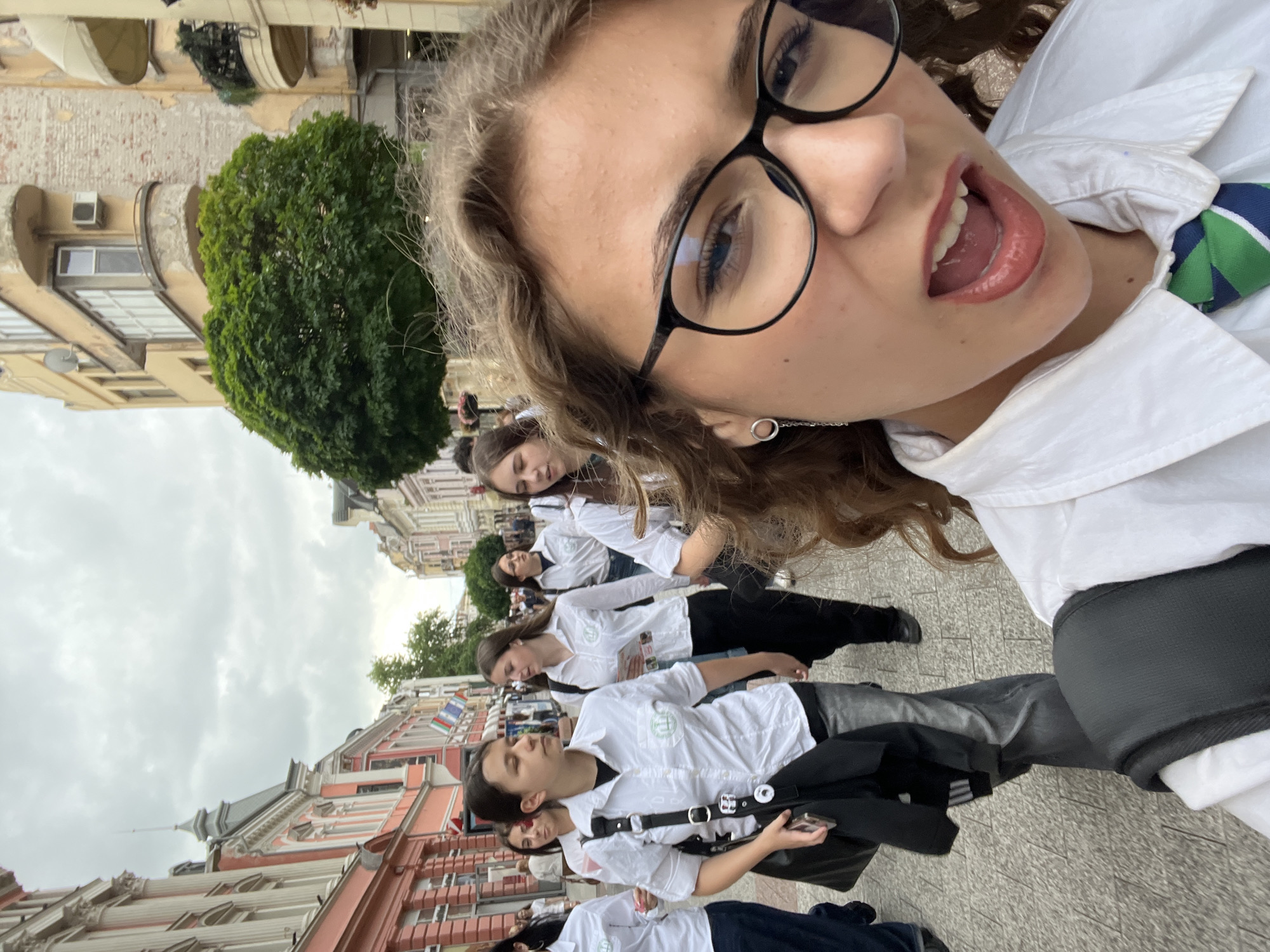 The image is a rotated selfie, capturing a lively urban street scene. The perspective is tilted 90 degrees clockwise, making the horizon appear vertical. In the immediate foreground, largely occupying the top right portion of the frame, is the face of a young woman taking the selfie. She has brown, curly hair, blue eyes, and wears black-rimmed glasses and a white button-up shirt with a green and blue striped tie. Her expression is playful, with her mouth open and tongue slightly extended. A silver hoop earring is visible on her left ear.
Behind her, a line of several other young women extends down the "street," which appears horizontally across the image. They are uniformly dressed in white long-sleeved button-up shirts, many featuring a small circular green logo on the left chest area, and dark pants or skirts. Their expressions are generally neutral, with most looking forward. They appear to be part of an organized group, possibly students on an outing.
The setting is an old city street, characterized by multi-story buildings with traditional European architecture, including ornate details, balconies, and some with awnings. The buildings exhibit varying pastel and earthy tones. A large, dense green tree stands prominently on the left side of the "vertical street" (middle distance). The ground is paved with tiles or cobblestones. The sky is overcast, indicating a cloudy daytime. On a distant building, a flag-like banner with green, red, and blue vertical stripes is visible. No specific readable text is discernible beyond the shirt logos.