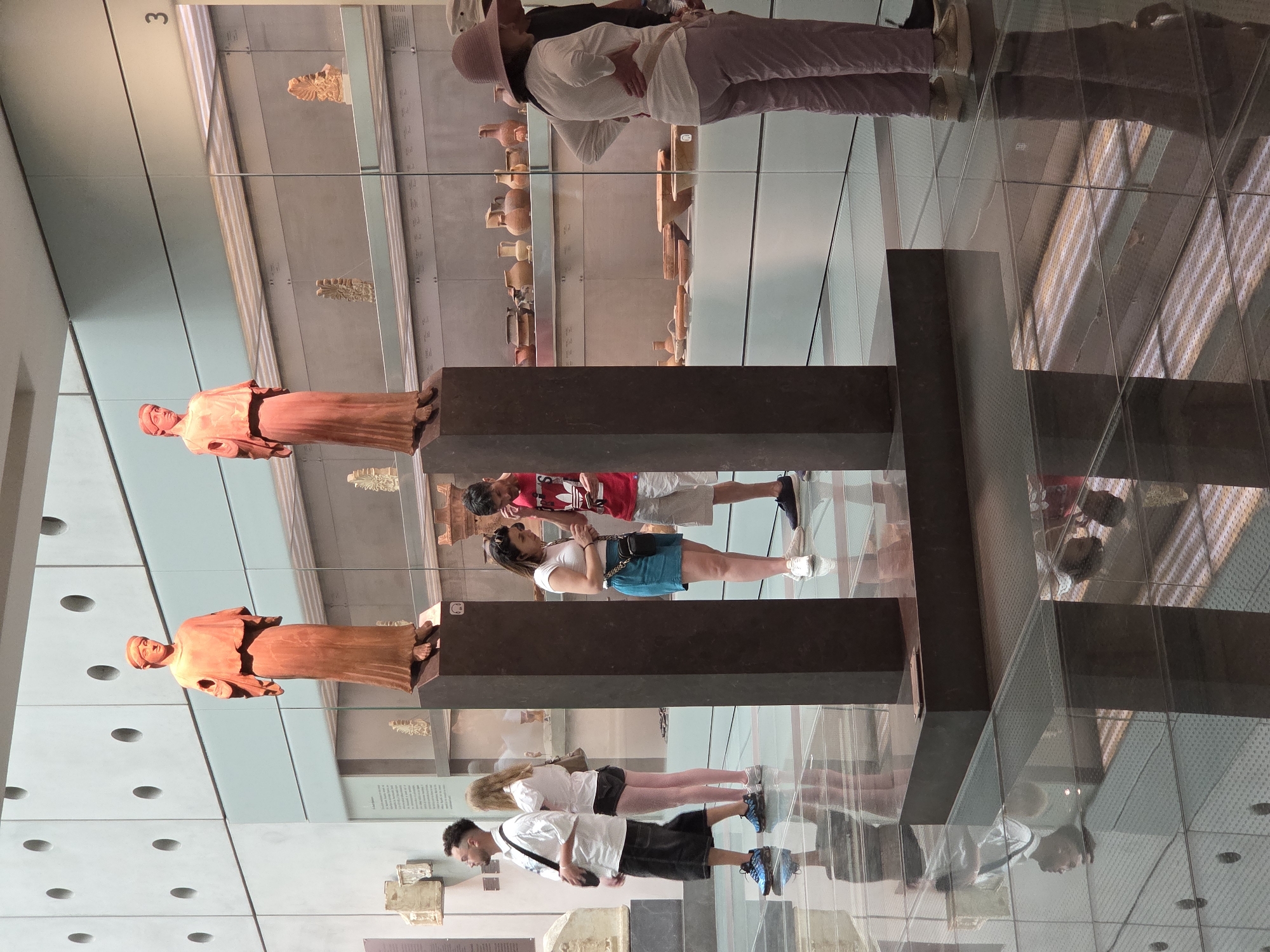 This is an interior shot of the Acropolis Museum in Athens, Greece. Two ancient Greek statues, a male and a female figure, stand on tall, dark pedestals in the foreground. The statues are reddish-orange in color and are depicted in flowing garments.
In the middle ground, a group of museum visitors are observing the exhibits. On the left, a man in a white shirt and black shorts is standing with his back to the camera. Next to him, a woman in a white shirt and black shorts is looking at the statue on the left. In the center, a woman in a white shirt and blue shorts is looking at her phone. To her right, a man in a red t-shirt and grey shorts is also looking at his phone. Further to the right, a woman in a white long-sleeved shirt and light purple pants is wearing a wide-brimmed hat and looking into a display case.
The background features glass display cases filled with ancient pottery and artifacts. The floor is made of reflective glass, creating mirrored images of the people and the statues. The overall lighting is bright and modern, typical of a museum environment. The number "3" is visible on a display case in the upper right corner.