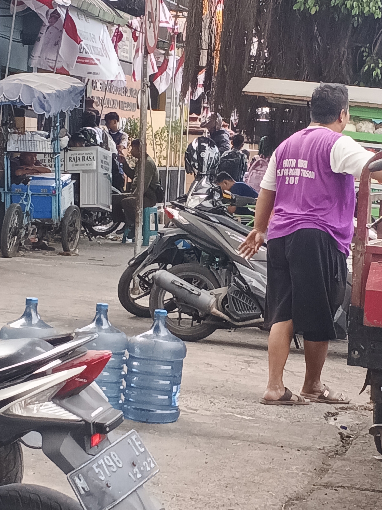 The image depicts a street scene in Krian, Indonesia, likely a market or a roadside area. In the foreground, several large blue water jugs are visible, some partially obscured by a motorcycle. To the right, a person wearing a purple vest over a white t-shirt and black shorts is standing, facing away from the camera. The purple vest has text printed on it, which appears to read "PANITIA BISA SJ. ROKHAN TESOR 202" and possibly more text below. The person is wearing brown sandals. Behind this individual, a motorcycle is parked. Further in the background, a blue and white food cart with a striped awning is set up, with several people gathered around it. Various red and white flags, possibly for a local event or celebration, are also visible, hanging from poles and structures. Another motorcycle is parked to the left of the food cart. The environment suggests an outdoor setting with paved ground and some vegetation in the upper right corner.