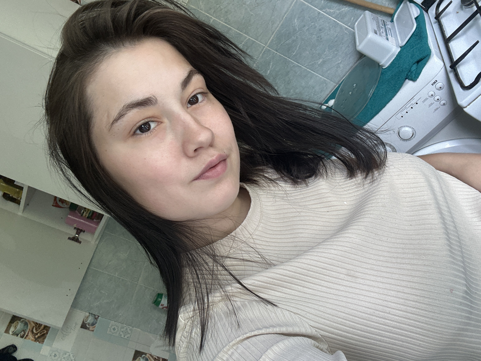 The image is a selfie taken indoors, likely in a kitchen or laundry area. The foreground features a young woman with dark hair, wearing a ribbed, cream-colored t-shirt. She is looking at the camera with a neutral expression. Behind her, a white washing machine is visible, topped with a teal towel and two white plastic containers. To the right of the washing machine, part of a white gas stove is in view. The background shows a tiled wall with a light blue-green tile pattern and a section of a kitchen cabinet. The overall scene suggests a domestic setting. There is no information in the image that allows for the identification of the city as Chaikovsky, Russia.