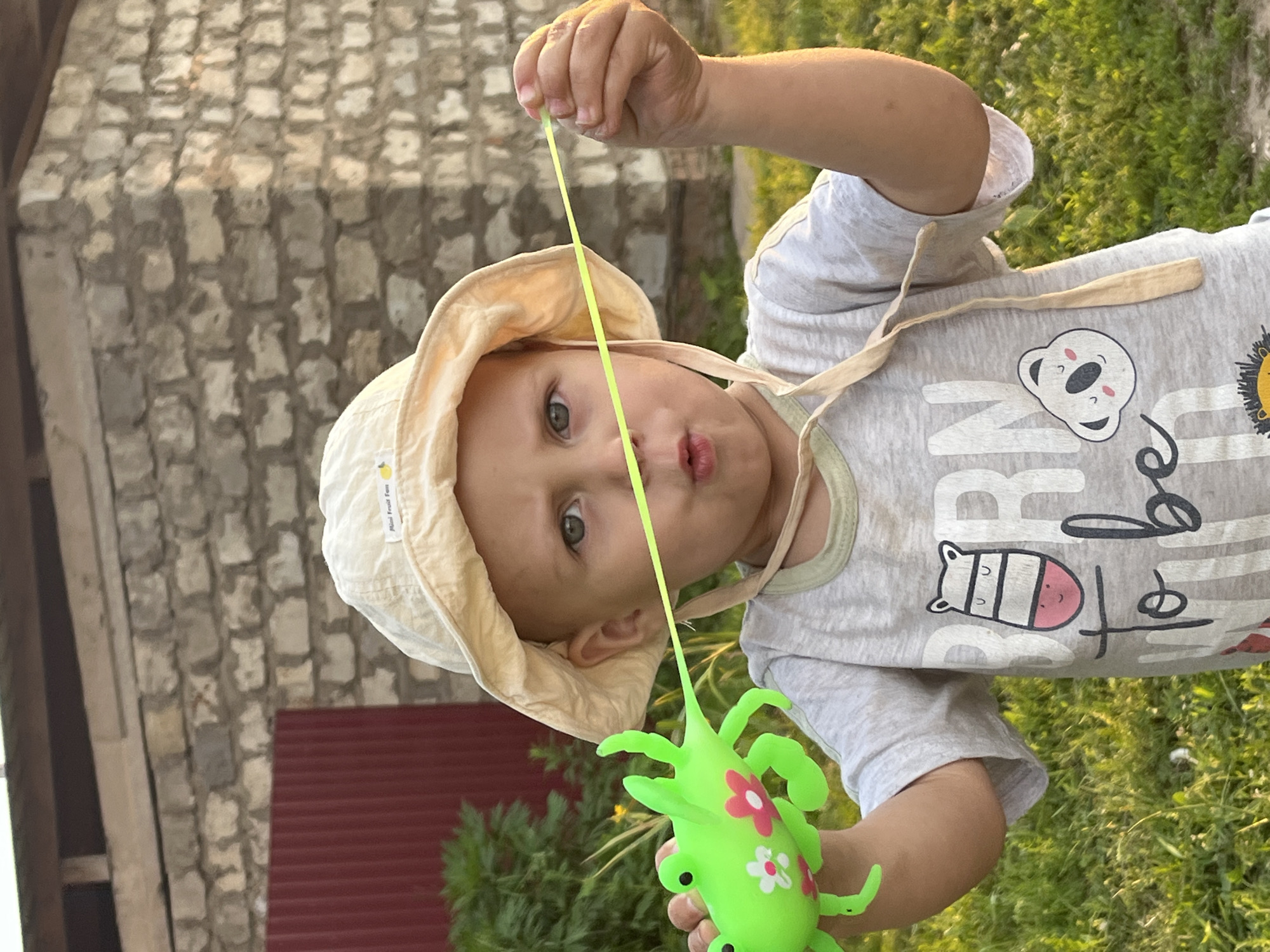 A young child wearing a light-colored bucket hat and a grey t-shirt is standing outdoors, holding a bright green toy frog with a yellow string attached. The child's mouth is puckered as they hold the string. In the background, there is a brick building and some greenery. The overall scene suggests a casual, outdoor setting, possibly a backyard in the city of Фоки, Russia.