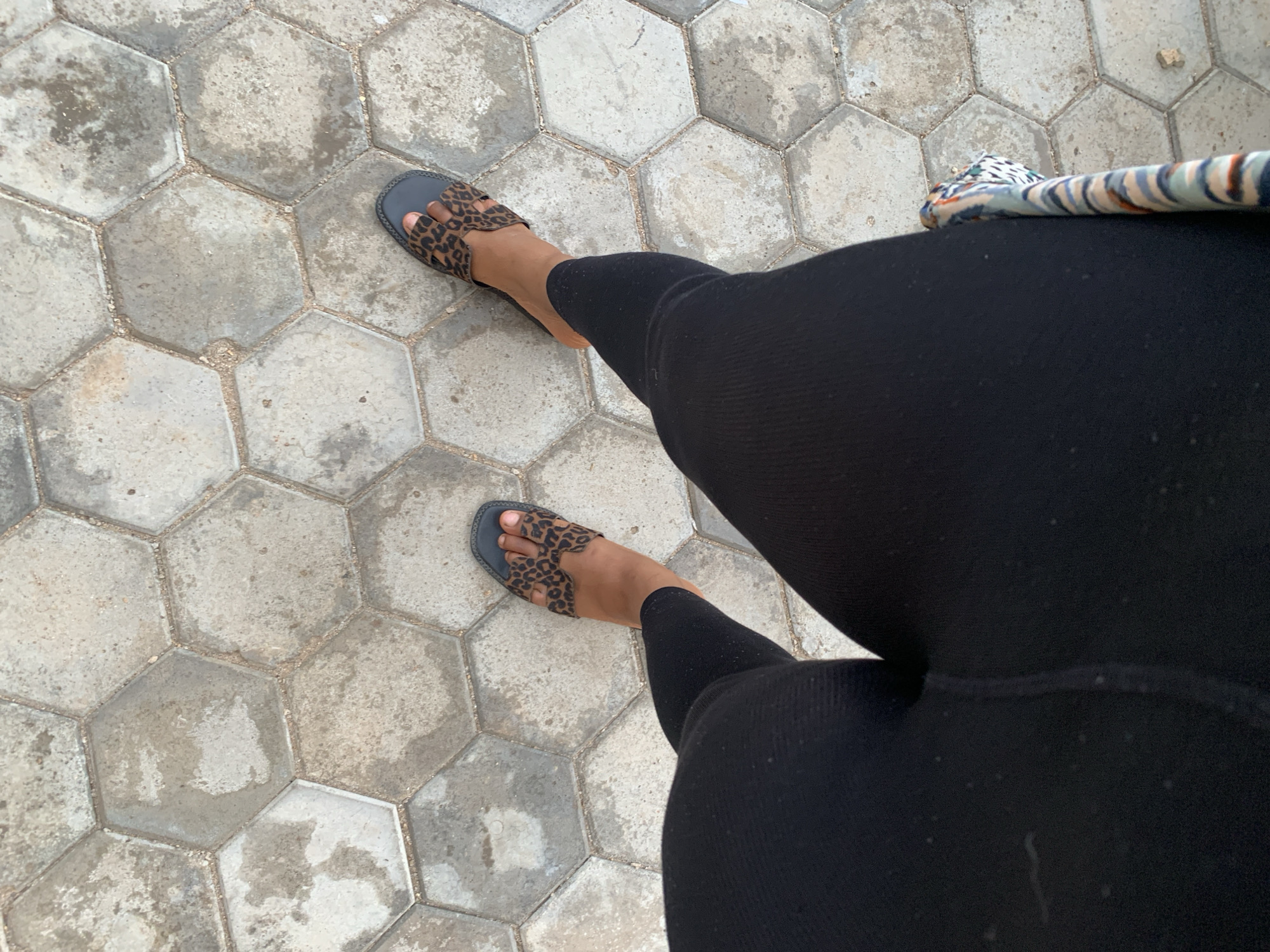 A woman wearing black leggings and leopard-print sandals stands on a hexagonal paver surface. A striped textile item is partially visible. The location is in a city, likely Jos, Nigeria, based on the user's prompt.
