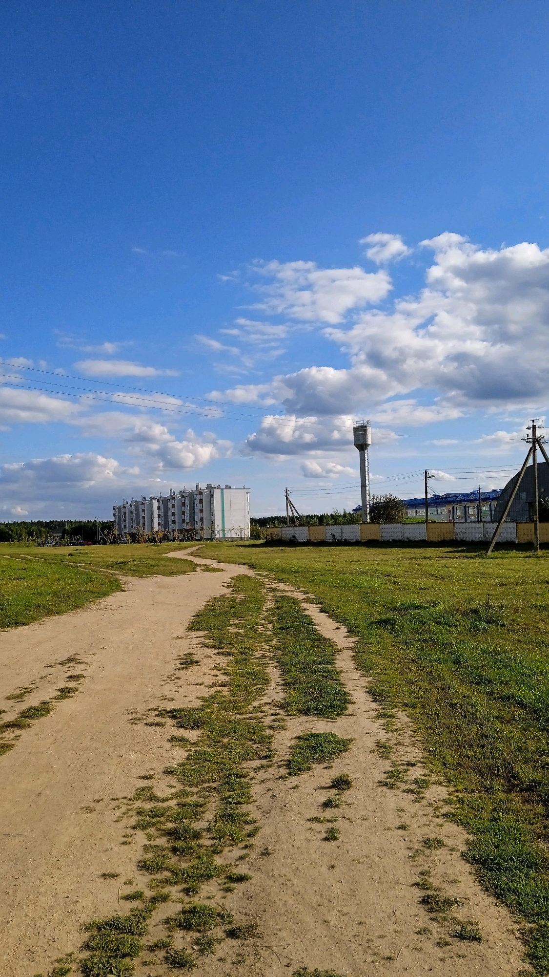 A dirt road cuts through a grassy field in a rural area, leading towards a cluster of apartment buildings and industrial structures. The sky above is a vibrant blue, dotted with fluffy white clouds. To the left, several identical apartment buildings with white and light green facades stand in a row, suggesting a residential development. Further to the right, a tall water tower rises above a long, blue-roofed building, possibly a factory or industrial complex, enclosed by a fence. Power lines crisscross the scene, supported by utility poles. The foreground is dominated by the dusty track, with patches of green grass growing through the compacted soil, indicating a well-traveled path. The overall impression is one of a developing rural landscape, blending residential and industrial elements with the natural environment under a clear, cloud-filled sky.