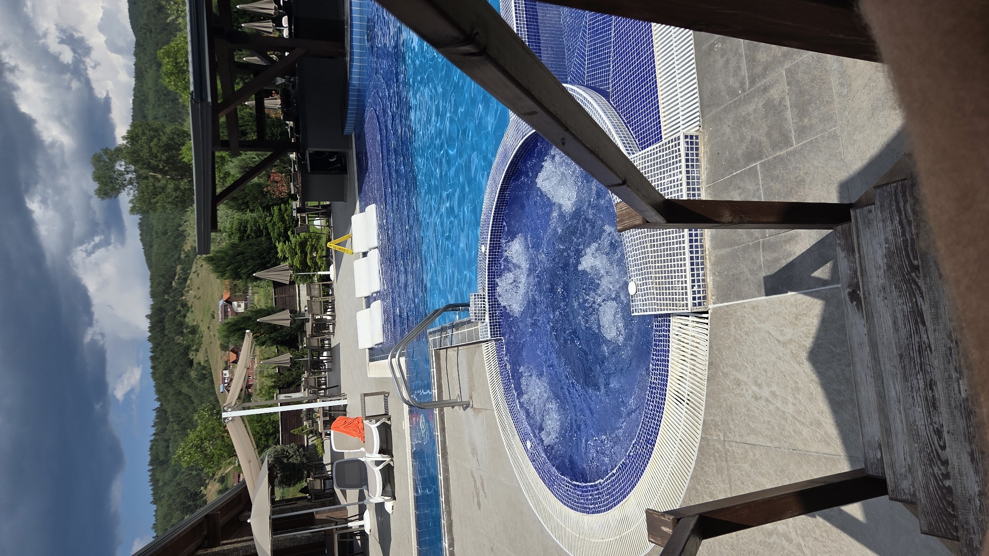 The image depicts an outdoor swimming pool area with a separate jacuzzi. In the foreground, a wooden staircase with dark brown railings leads down into the scene. The jacuzzi is circular, tiled with blue and white mosaic tiles, and filled with bubbling blue water. Adjacent to the jacuzzi is a larger swimming pool, also tiled in blue. Several white lounge chairs are arranged in a shallow section of the pool. In the background, there is a patio area with tables and chairs, some covered by beige umbrellas. Beyond the patio, a building with a sloped roof is visible, as is a tree-covered hillside with scattered houses. The sky is a mix of dark clouds and patches of blue. The overall environment suggests a resort or recreational facility. No people are clearly visible in the image, but a shadow of a person is cast on the tiled ground in the foreground.