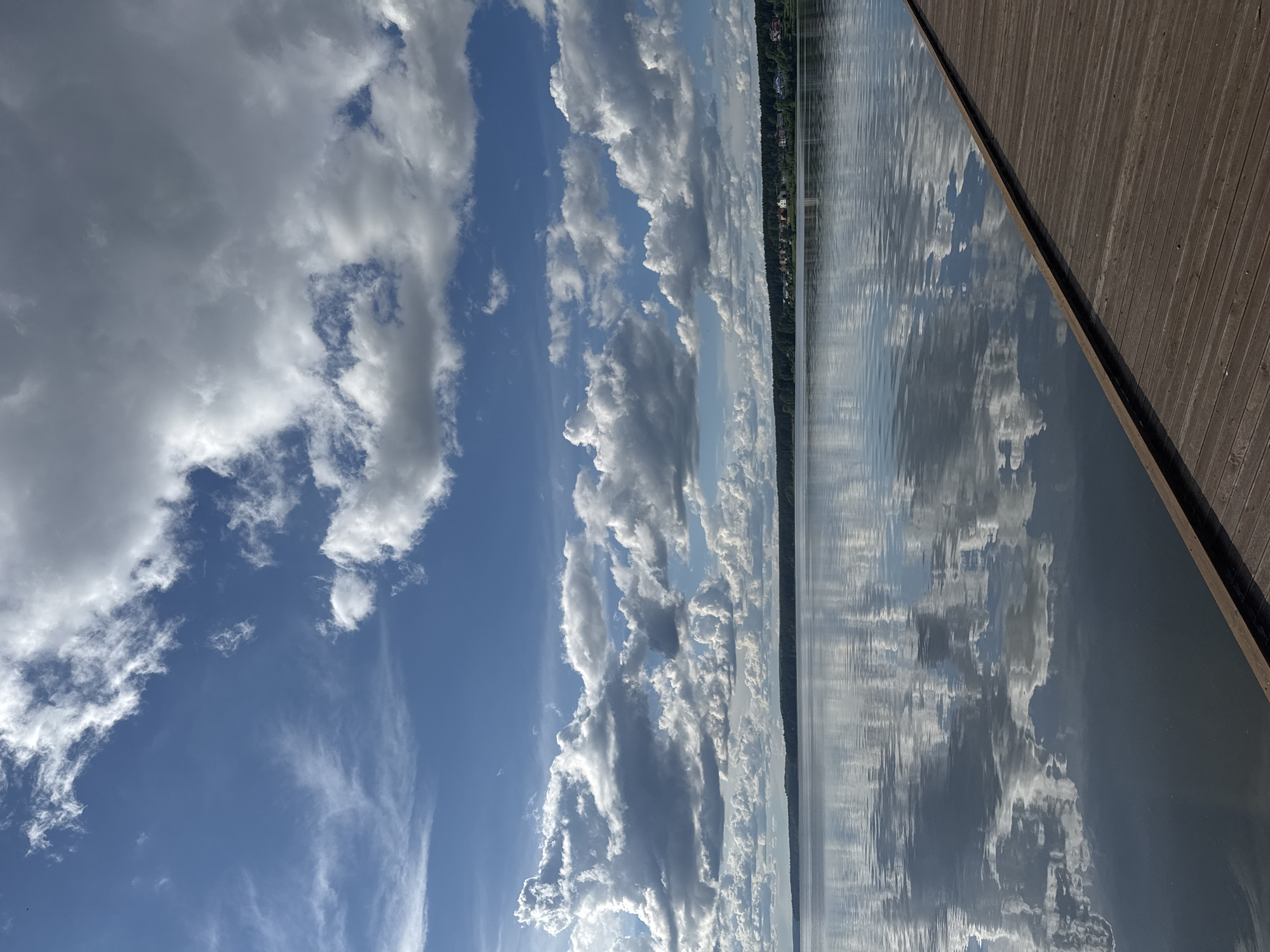 The image shows a body of water reflecting a sky filled with cumulus clouds. A strip of land with trees and buildings is visible in the distance. A wooden dock is present on the right side of the frame. The scene appears to be located in a natural environment. Without more information, it is difficult to confirm that the location is Votkinsk, Russia. However, the presence of a body of water and a forested shoreline is consistent with the geography of Votkinsk.