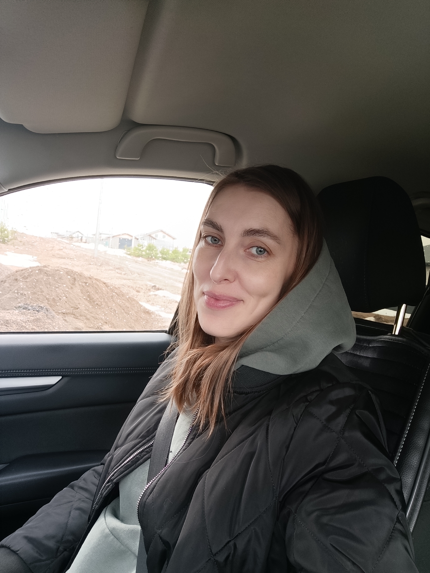 A woman is seated in the driver's seat of a car. She is wearing a black quilted jacket and a gray hooded sweatshirt. A seatbelt is fastened across her chest. The car's interior is black leather and plastic. The woman is looking toward the camera with a slight smile. Outside the car window, there is a construction site with piles of dirt and a few small buildings in the distance. This could possibly be in Chaikovsky, Russia.