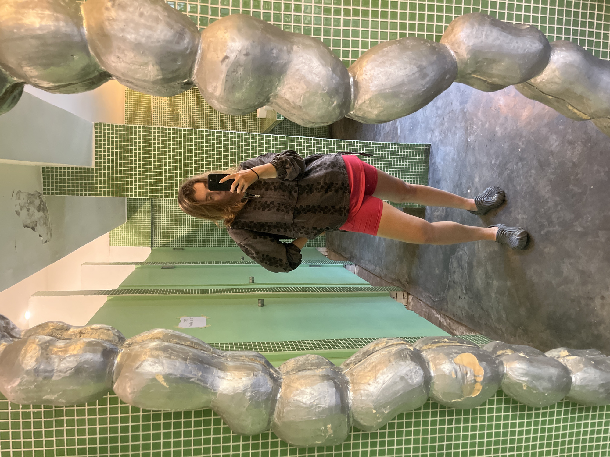 A person is taking a mirror selfie in a bathroom. The bathroom has green tile walls and gray tile floors. There are green bathroom stalls visible in the reflection of the mirror. The mirror has a silver, bulbous frame. The person in the reflection is wearing a gray and black zip-up sweatshirt and red shorts, and gray shoes.
The image was taken in North Kuta, Indonesia.