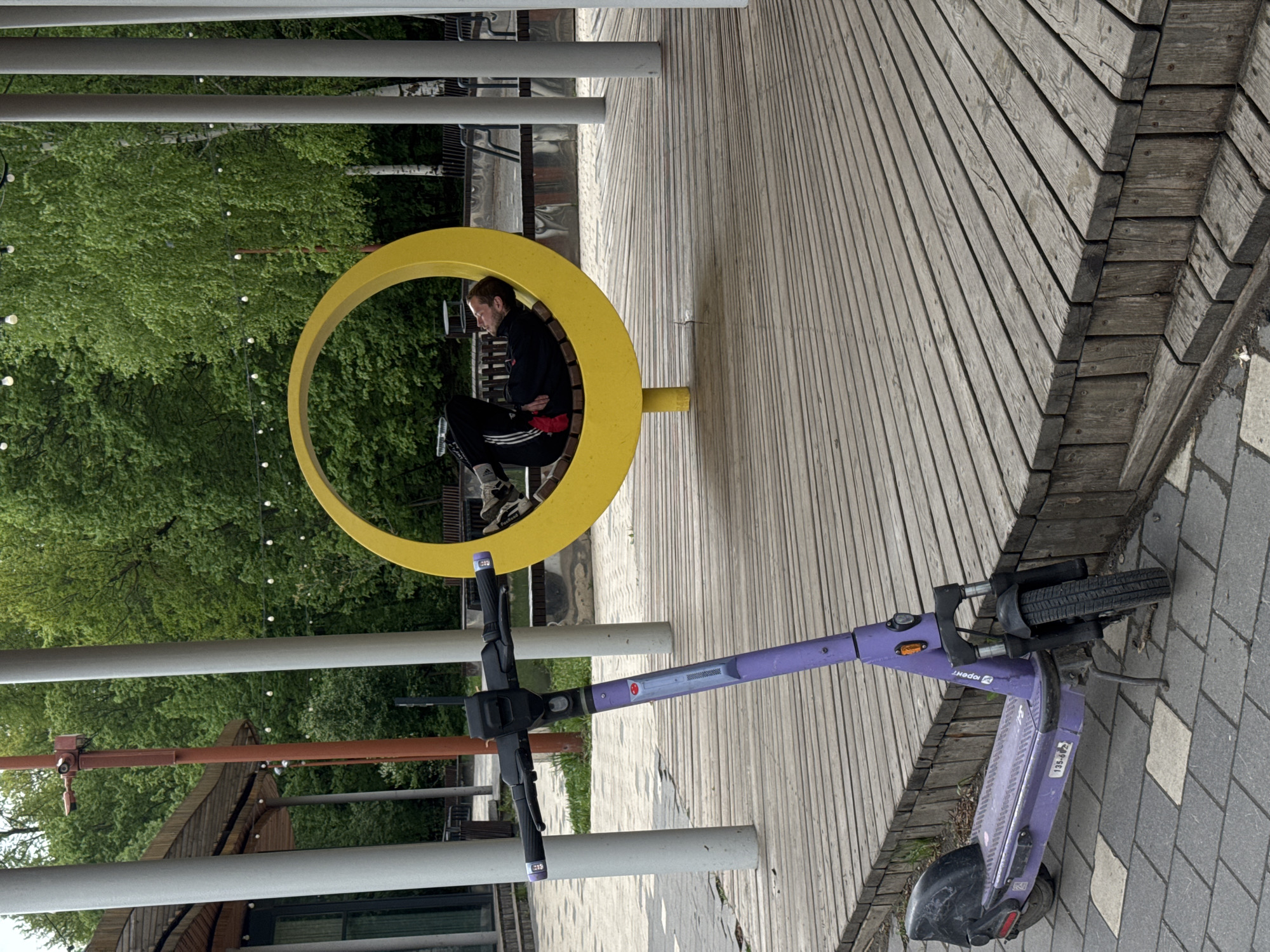 A medium shot captures a scene in what appears to be a public space in Kazan, Russia. A man sits in a yellow, circular bench, dressed in a black jacket and track pants. He seems to be engrossed in thought. A parked purple electric scooter lies horizontally on the right side of the frame, partially on a wooden boardwalk and partially on a paved area. The boardwalk is made of light-colored, parallel wooden planks, while the paved area consists of gray bricks. The backdrop shows lush green trees, indicating the location is near a park or green space. Metal posts and beams overhead are visible, suggesting an outdoor structure like a pergola or covered walkway.