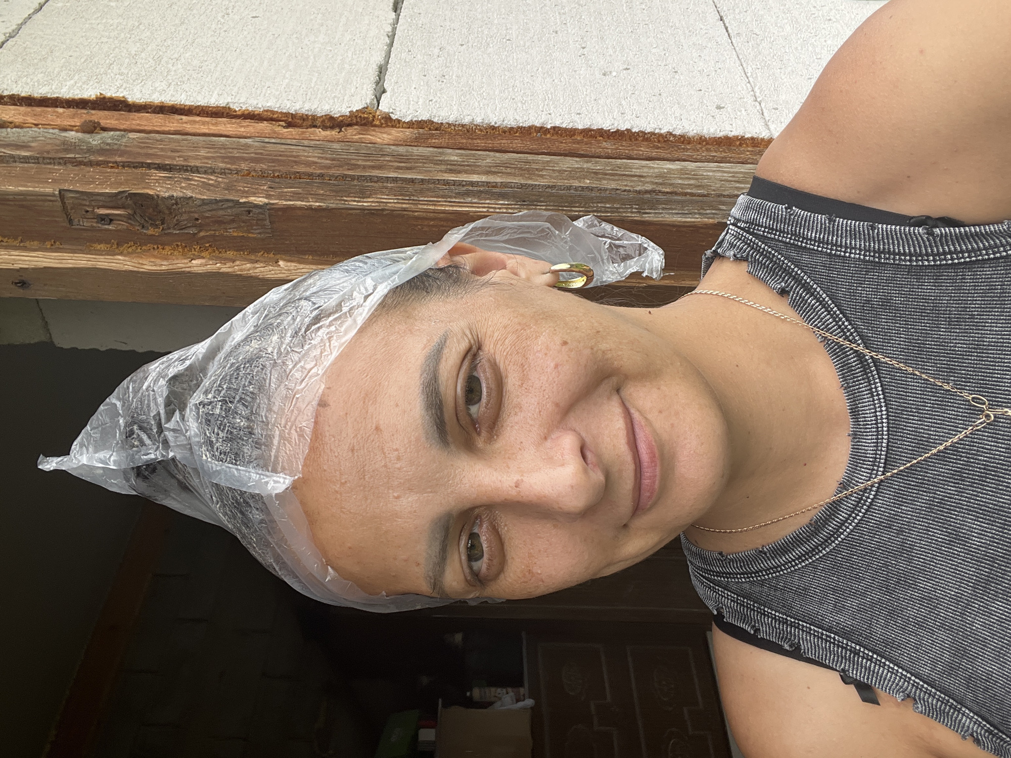 A woman with medium-toned skin and dark hair is shown from the chest up, outdoors, with a plastic bag covering her head like a makeshift shower cap. She is wearing a grey tank top with frayed edges and a thin gold necklace. Her expression is a slight smile, and her eyes are open. The background shows a rustic wooden door frame and a white concrete wall. The scene appears to be in a residential area in Andreyevo, Russia, suggesting a casual, perhaps domestic, setting.