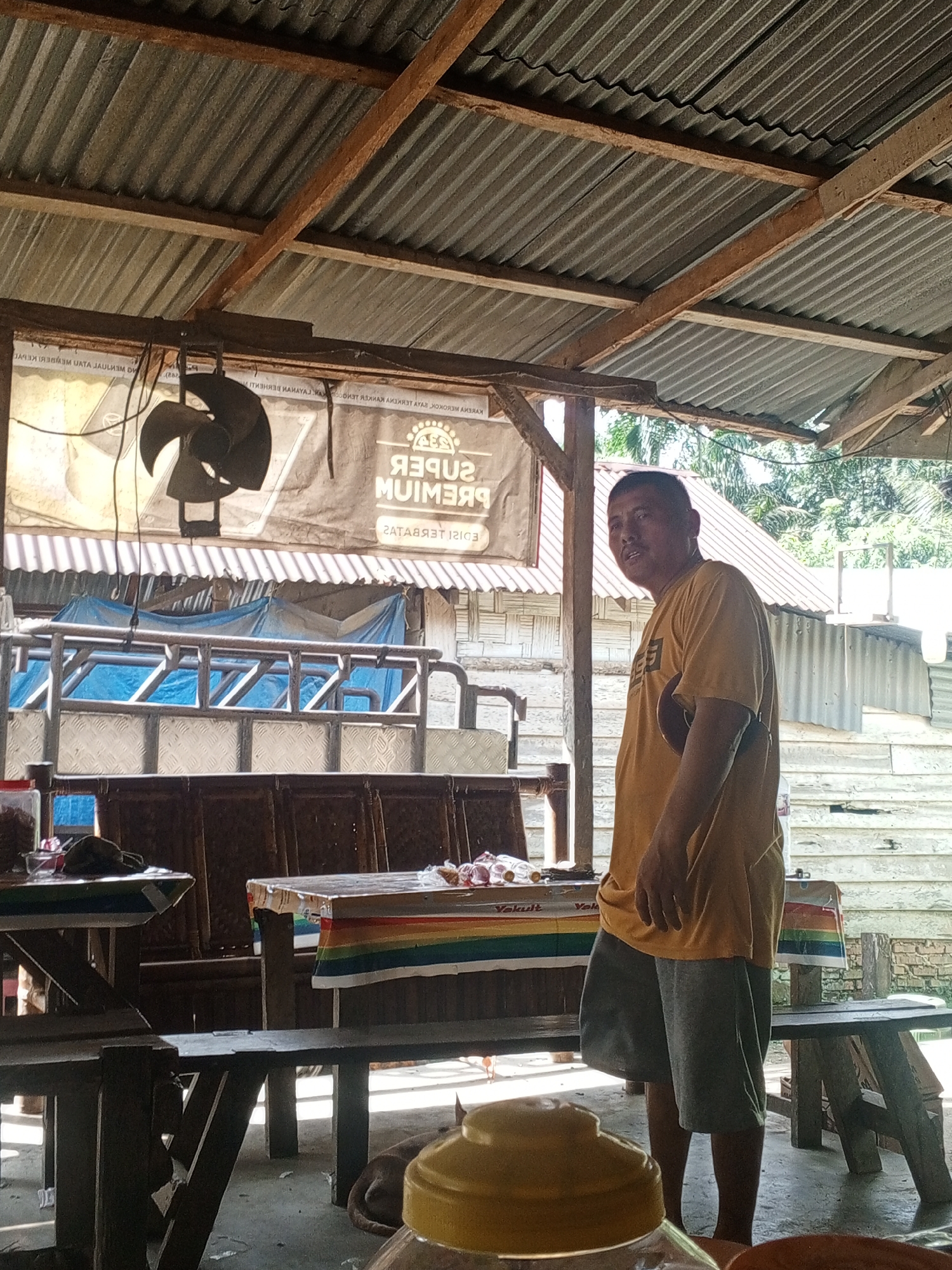 A man in a yellow t-shirt and grey shorts stands in an open-air shelter with a corrugated metal roof supported by wooden beams. A fan is suspended from the ceiling, and a banner with text, including "SUPER PREMIUM," is visible behind him. In front of him are wooden tables covered with colorful cloths. A truck bed with blue tarpaulin is visible behind the tables. The environment appears to be rural, with trees and buildings in the background. This scene is likely located in Tanah Gara Hulu, Indonesia, given the details of the structure and surroundings.