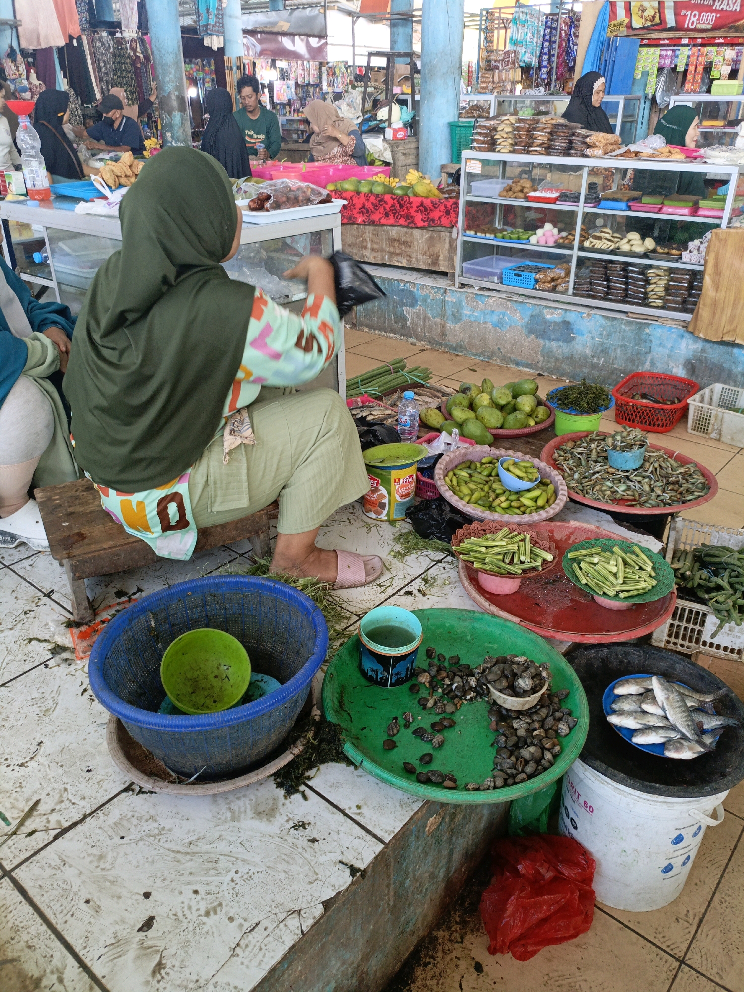 The image depicts a street market scene in Bulukumba, Indonesia, likely a traditional wet market. In the foreground, a woman wearing a green hijab and a patterned top is seated on a wooden stool. She is holding a black plastic bag, suggesting she is a vendor or a shopper involved in a transaction. Around her, the ground is tiled and somewhat soiled, with various produce and seafood displayed in baskets and trays. These include green okra, avocado-like fruits, small fish, and shellfish. Several individuals, some also wearing hijabs, are visible in the background, browsing stalls that sell packaged goods and food items. The environment appears bustling with commercial activity.