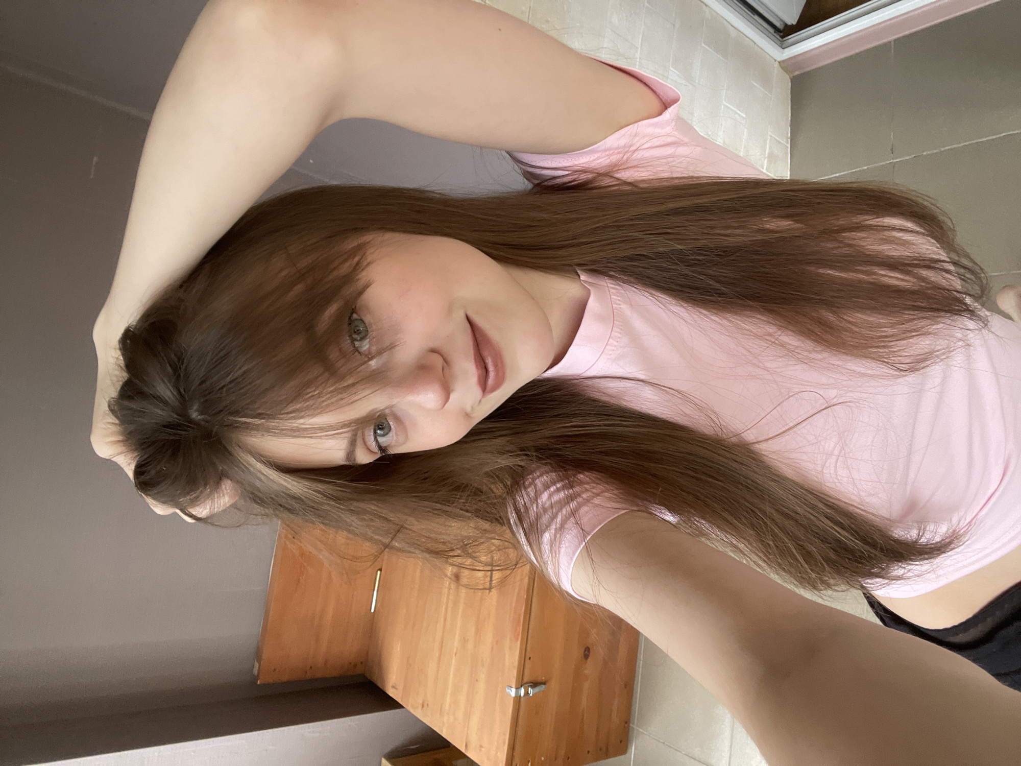 A woman with long brown hair and fair skin is posing for a selfie. She is wearing a pink t-shirt and black shorts. Her left arm is raised above her head, while she holds the camera with her right hand. A light brown wooden cabinet with silver handles is visible behind her. The background includes a gray wall and a tiled floor. The location is potentially a private residence, possibly in Ufa, Russia, but there is no definitive evidence of a specific city or country in the image.