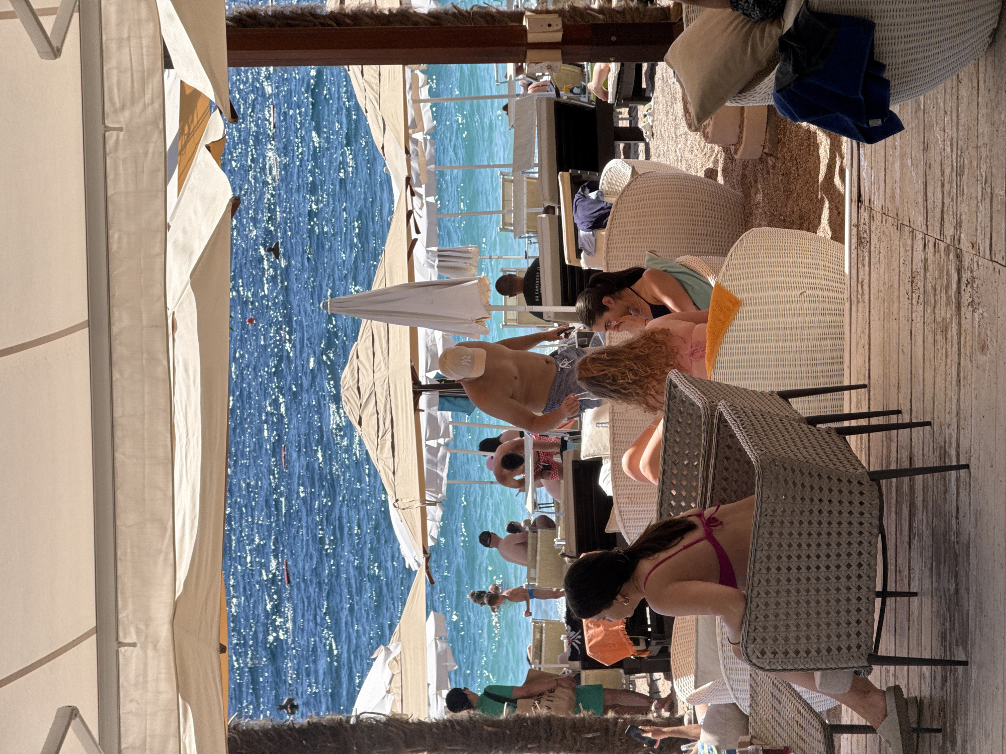 In Kamenovo, Montenegro, a beachside cafe is captured during daylight. The foreground features patrons seated at wicker furniture, with a woman in a pink bikini visible in the left chair. Beyond the cafe, the turquoise Adriatic Sea sparkles under the sun, with several beach umbrellas and lounge chairs set up on the sandy shore. People are seen swimming and relaxing by the water. The scene is characterized by a relaxed, summery atmosphere, indicative of a popular tourist destination.
