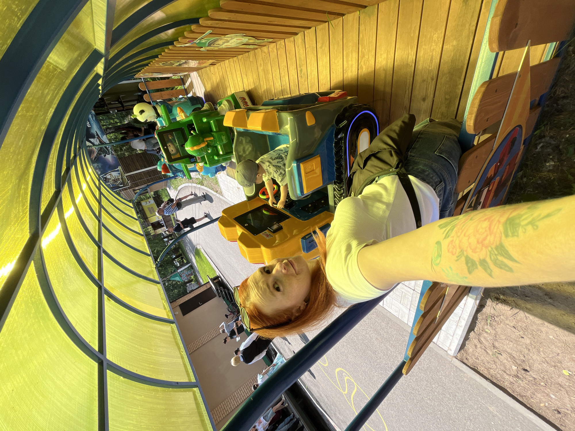 This image captures a scene at the Chaikovsky City Park in Chaikovsky, Russia, focusing on a children's amusement ride. In the foreground, a woman with red hair and a white t-shirt is taking a selfie. She has tattoos on her arm. Behind her, a yellow and blue tunnel-like structure with a translucent yellow roof arches over the ride. The ride itself consists of several colorful, cartoon-themed vehicles. One vehicle, a gray and blue jeep-like car, is occupied by a young child wearing a grey cap and a patterned t-shirt, who appears to be steering. Another green, robotic-themed vehicle is visible behind it. The ride is on a wooden platform adjacent to a paved area. In the background, other park-goers are visible, along with trees and a building. The overall atmosphere suggests a sunny day at an amusement park.
