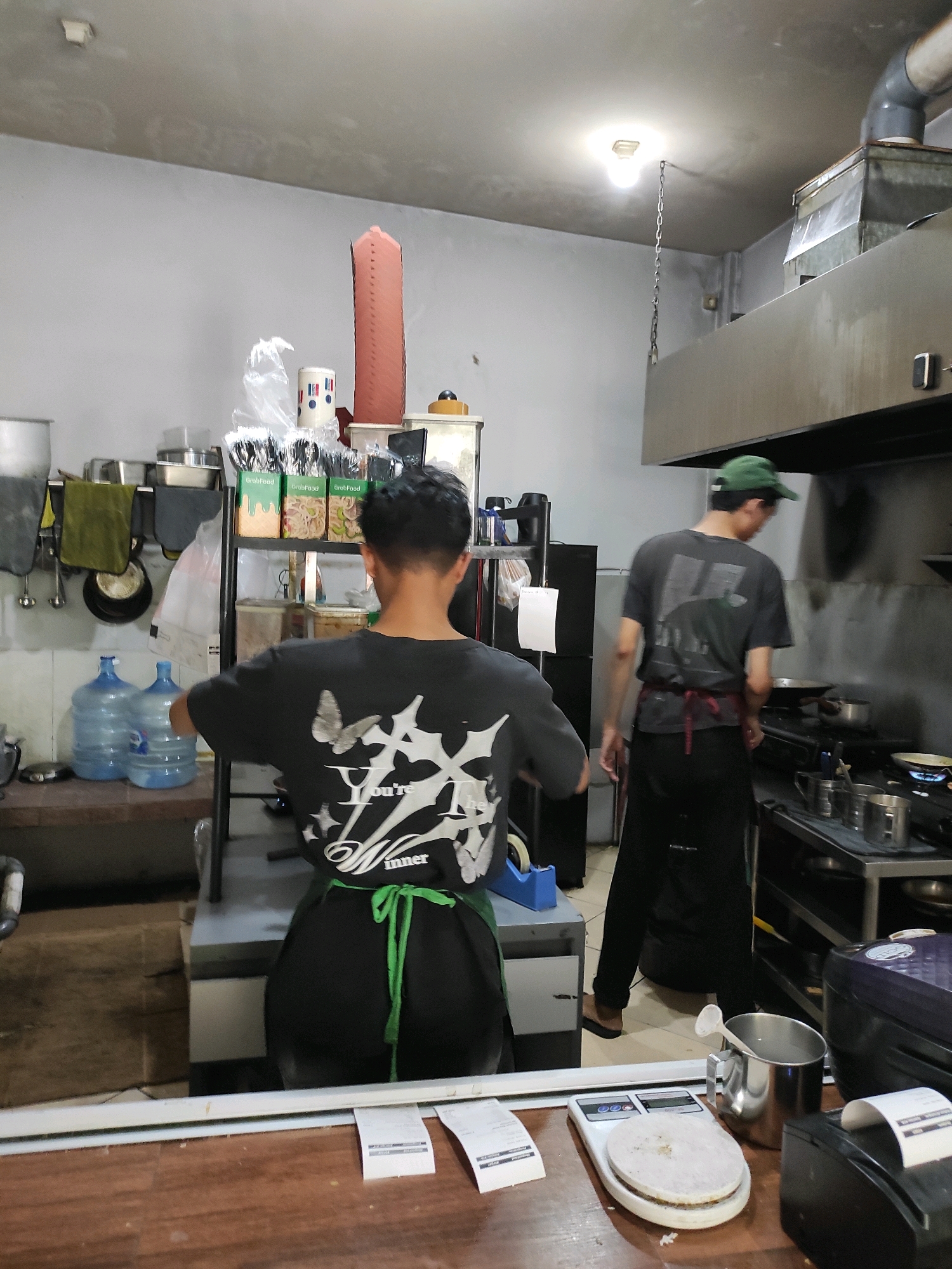 In a commercial kitchen located in South Jakarta, Indonesia, two individuals are present. The foreground is dominated by a wooden counter with receipts and a digital scale. Behind the counter, a young man wearing a black t-shirt with a butterfly design and a green apron is seen from the back, appearing to be working. Further back, another man, also viewed from behind, wears a dark t-shirt, a green cap, and a dark apron, and is standing near a stove. The kitchen area is equipped with stainless steel appliances, ventilation systems, shelves stocked with food items, and large water dispensers. The overall scene depicts a functional food preparation environment.