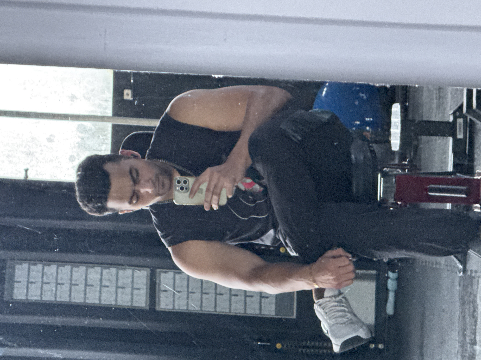 A young man is captured in a mirror selfie, seated on a piece of gym equipment. He is wearing a sleeveless black t-shirt and black pants, with white athletic shoes on his feet. His left leg is crossed over his right, and he holds a light green smartphone in his right hand, using it to take the photo. He appears to be wearing an earbud in his left ear and a chain around his neck. A thin gold bracelet is visible on his right wrist. The background suggests a gym environment with mirrors, weightlifting equipment, and what appear to be window panes or frosted glass panels. The image does not contain any information that would allow for its location to be identified as Kolkata, India.