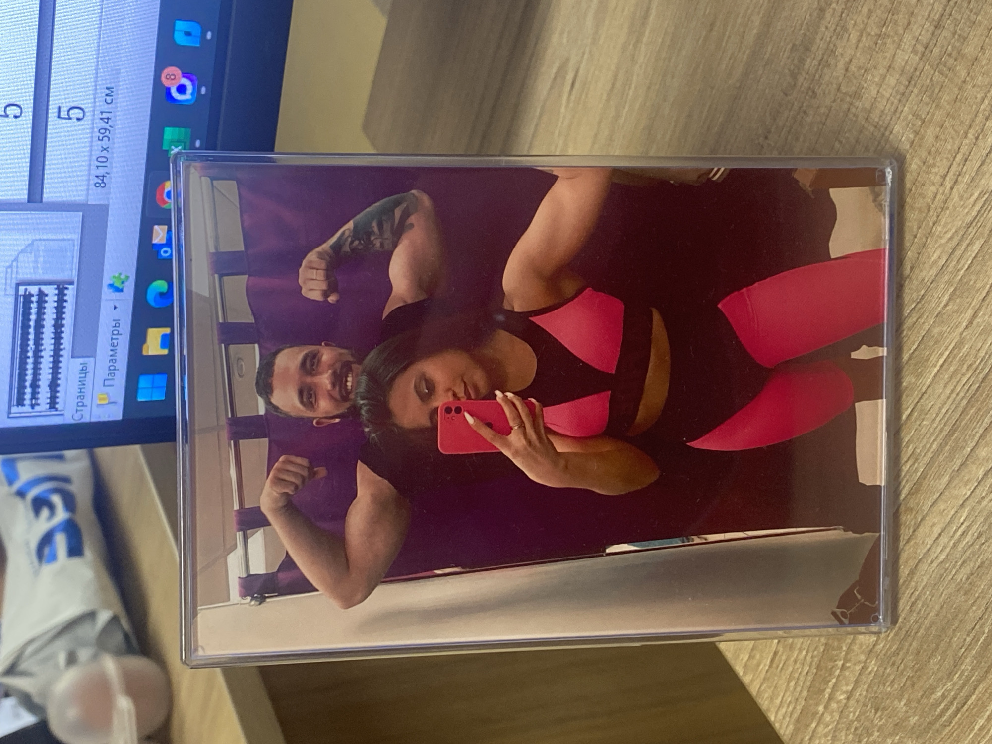 A photograph in a clear plastic frame shows a man and woman in workout clothes posing for a selfie in front of a purple background. The man is flexing his arm; he has a tattoo on his upper arm. The woman is holding a red phone and taking the selfie. The photograph and a laptop are resting on a wooden surface. The laptop displays icons and software windows. A white package with blue lettering is visible. The location is not discernible from the image. No evidence indicates the city of Luzhitsy, Russia.