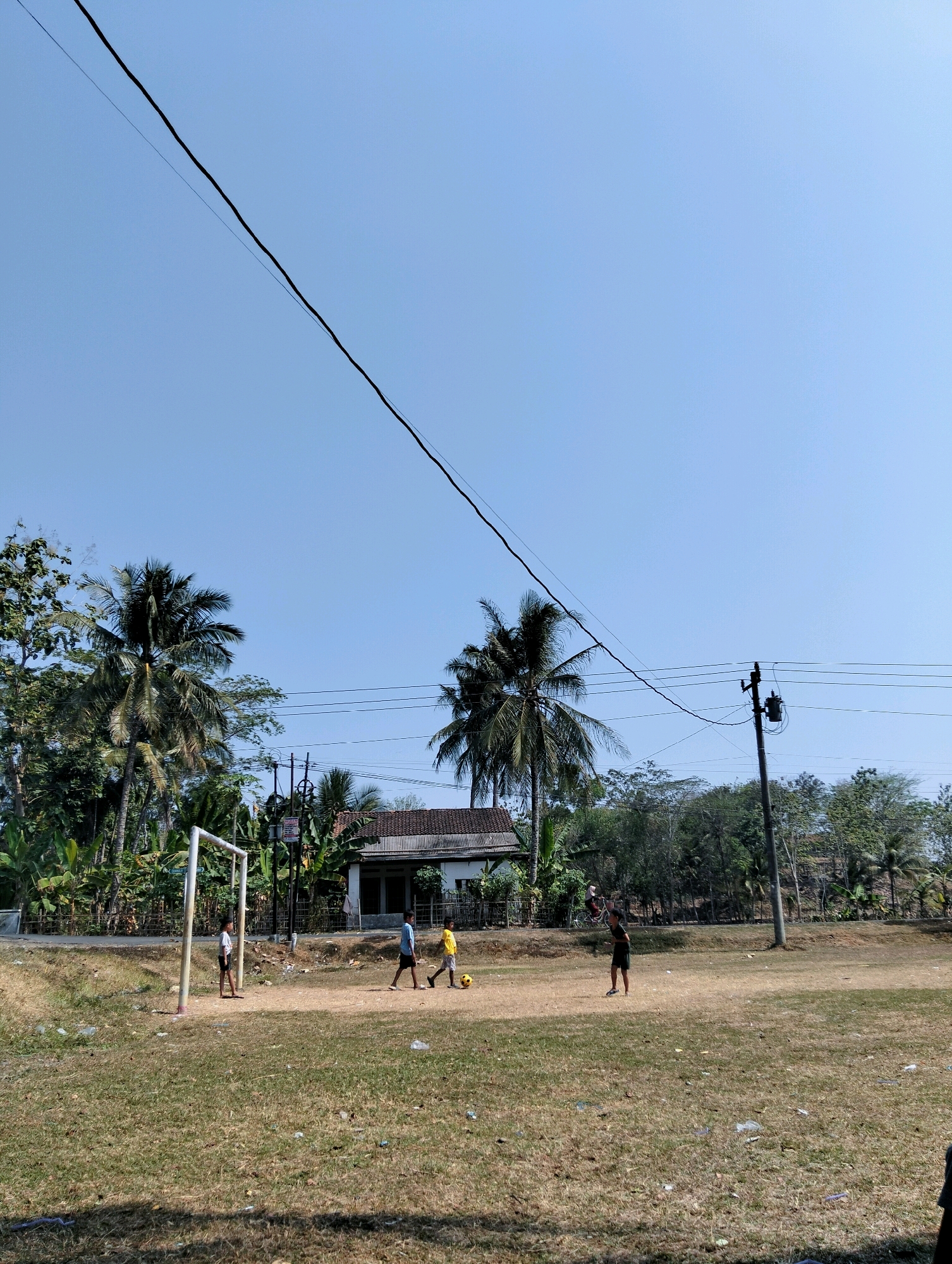 The image depicts an outdoor scene in Tipar, Indonesia, featuring a dirt field with sparse, dry grass. A makeshift soccer goal stands on the left side of the frame. Three young boys are visible on the field, engaged in a soccer game. One boy in a white t-shirt stands near the goal, while two others in a blue and yellow shirt respectively are playing with a soccer ball. In the background, a small house with a tiled roof is situated amidst lush vegetation, including several palm trees. Electrical poles with wires crisscross the sky, indicating the presence of infrastructure. The sky is clear and blue, suggesting a sunny day. The overall impression is of a casual, community sporting activity taking place in a rural or semi-rural setting in Indonesia.
