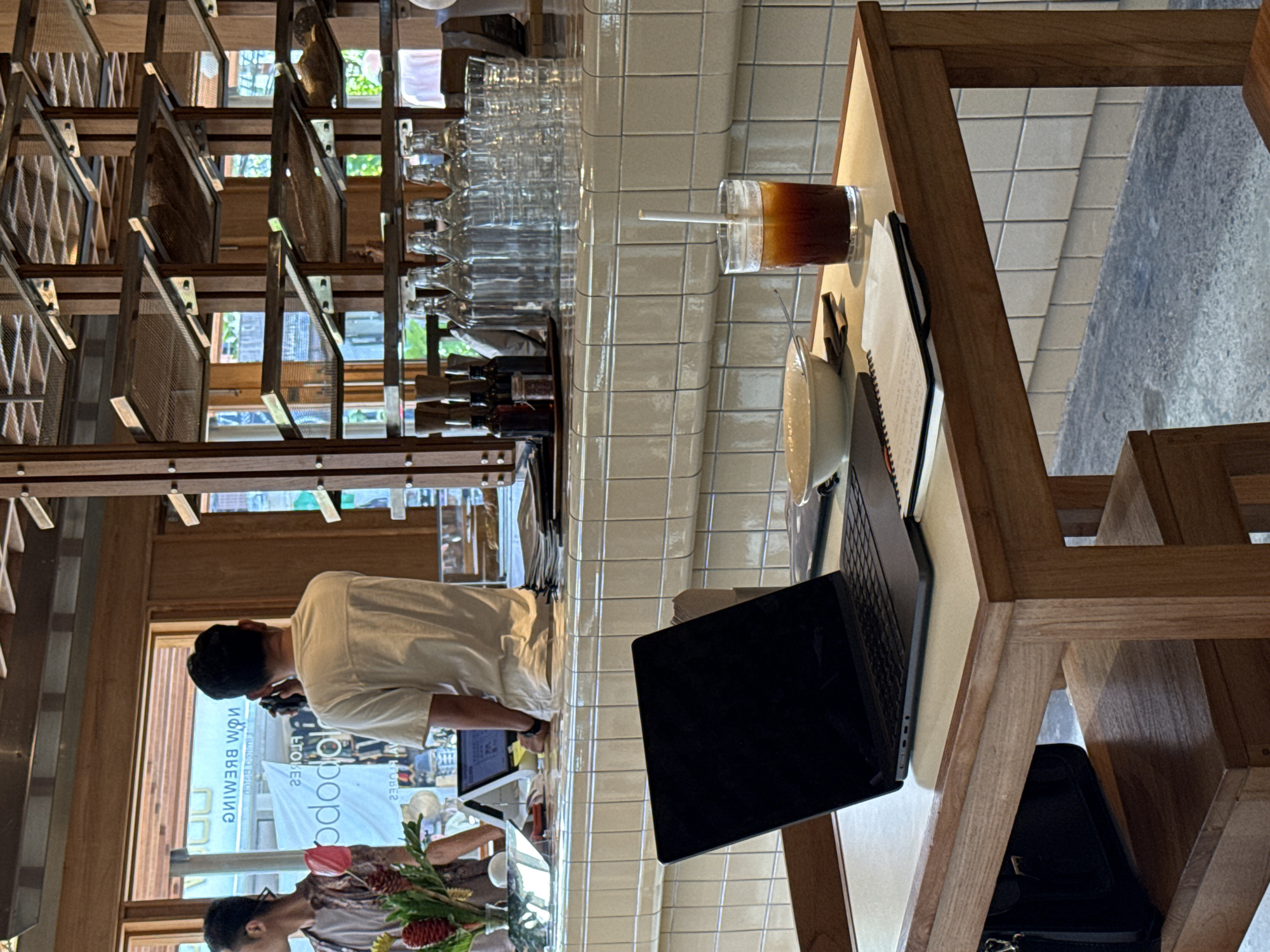 Here is a description of the image:
The image captures a coffee shop setting. A man wearing a white shirt is standing near a counter that has a POS system. A laptop is open on a table. There is a glass of coffee, a coffee mug, and a notebook, also on the table. A vase with flowers is placed near the counter. The coffee shop has a rack with glassware on the wall. A man can be seen through the window, which has the name "NOW BREWING" written in reverse. The POS system displays the name "podopo", also written in reverse. The style of tiling indicates that the location is likely in North Kuta, Indonesia. A black bag rests beneath the table, and the chairs are made of wood.
