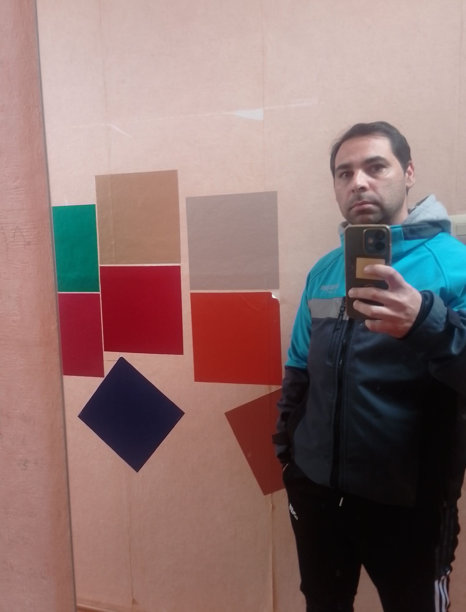 A man is taking a selfie in a room with a mirrored wall. The man is wearing a turquoise and black hooded jacket and black track pants with white stripes. He is holding a smartphone in his right hand. The mirrored wall reflects the man and also shows a series of colored squares arranged on a beige surface behind him. The colored squares include green, tan, gray, red, orange, and dark blue. The room appears to be an indoor space, possibly a studio or a room with a display of materials. There is no specific indication of the city of Delmenhorst, Germany.
