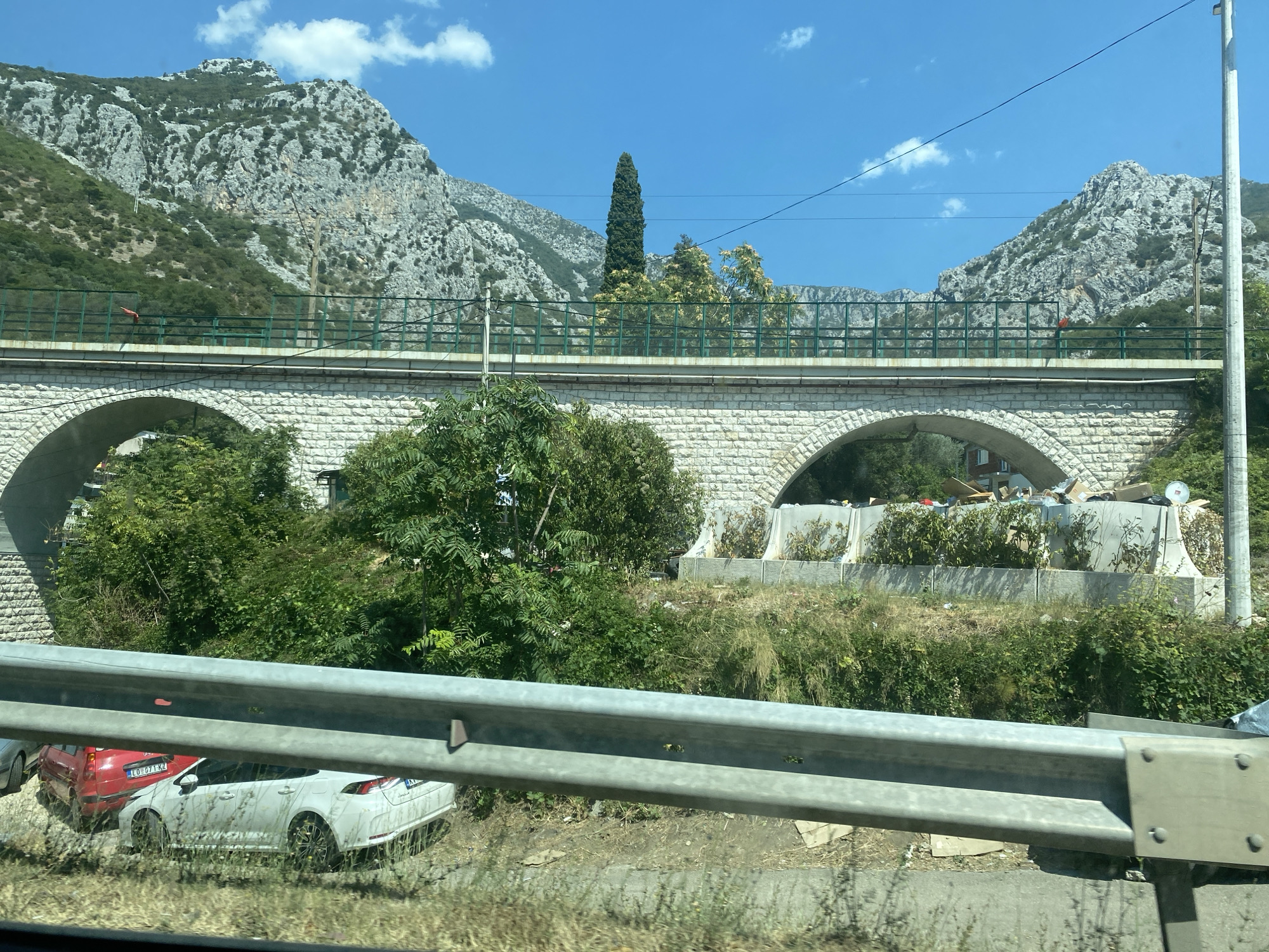 The image captures a view of a stone arch bridge spanning over an area with dense vegetation and a road. The bridge's arches are visible, and a green metal railing runs along its top. Behind the bridge, a rugged, mountainous landscape dominates the background, with rocky slopes and patches of greenery under a clear blue sky with scattered white clouds. In the foreground, a metal guardrail is prominently placed, with a part of a white car visible to the left.
The scene appears to be located in the vicinity of Sutomore, Montenegro, given the characteristic mountainous terrain and architectural style often found in the region. The bridge itself is an impressive structure, suggesting it might be part of a road or railway line that crosses the valley. The surrounding vegetation is lush, indicating a natural and possibly rural setting, though the presence of the bridge and cars hint at human infrastructure. The overall impression is one of a scenic yet functional landscape.