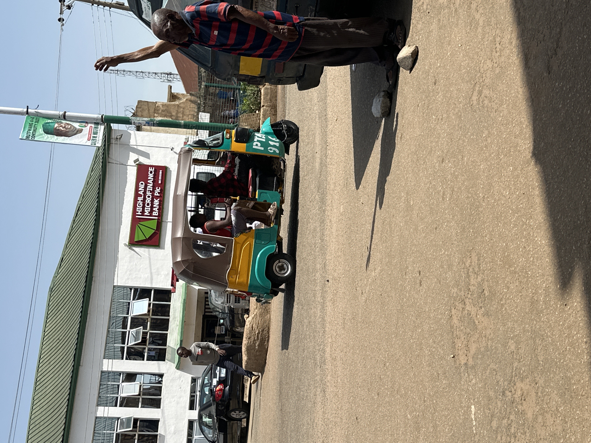 This image captures a street scene in Bukuru, Nigeria, on a sunny day. A bright green and yellow auto-rickshaw, identified with the letters "PTA" and the number "916" painted on its side, is prominently featured. It appears to be in motion or stopped at the side of the road. Two individuals are visible within the auto-rickshaw; one is driving, wearing a red and black checkered shirt, and the other, partially obscured, is wearing a white tank top.
To the right of the auto-rickshaw stands an elderly man, dressed in a striped blue and red shirt and dark pants, with his right arm raised. He is facing the auto-rickshaw, possibly interacting with its occupants or signaling. Two rocks are on the ground near his feet.
In the background, a white building with a green roof is visible. It has multiple windows, some of which are open. A sign on the building reads "HIGHLAND MICROFINANCE BANK PIC". A black sedan is parked to the left of the auto-rickshaw, with another dark-colored vehicle behind it. A young man in a grey long-sleeved shirt and dark pants is walking away from the camera, towards the building. A banner with a picture of a man in a green cap and the text "MUTFWANG 4+4 CONTINUITY 2023" is attached to a pole near the building. The environment suggests an urban street with moderate traffic and commercial activity.