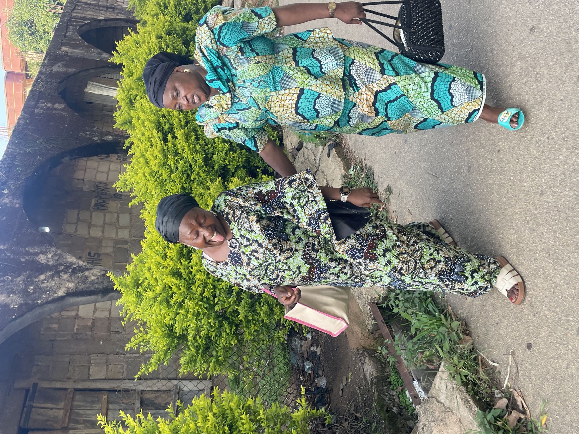 The image captures two women walking on a paved surface in the city of Jos, Nigeria. The woman on the left, wearing a black headwrap and a green, black, and white patterned traditional outfit, has her eyes closed, mouth open, and tongue extended in a joyful or playful expression. She carries a light-colored tote bag with pink trim on her left arm and wears light-colored sandals with red nail polish on her toes. The woman on the right, also wearing a black headwrap, is dressed in a teal, green, and gold patterned two-piece traditional outfit with an elaborate neckline. She looks towards the left with her mouth slightly open and carries a black woven bag in her left hand, wearing teal slide sandals.
Behind the women, a dense row of bright green bushes spans the mid-ground. Beyond these bushes, a weathered concrete block wall with multiple arched openings is visible. White graffiti reading "PROPERTY IS NOT FOR SALE BEWARE" (partially obscured) is spray-painted on this wall. To the far left, a wooden door or gate is partially visible behind the bushes and a chain-link fence. The ground beneath the women is grey pavement. To the immediate left of the pavement, a narrow strip of dirt contains sparse vegetation, discarded plastic debris, and a drainage channel partially covered by a rusty metal grate. Distant reddish-brown buildings and trees are visible above the wall.