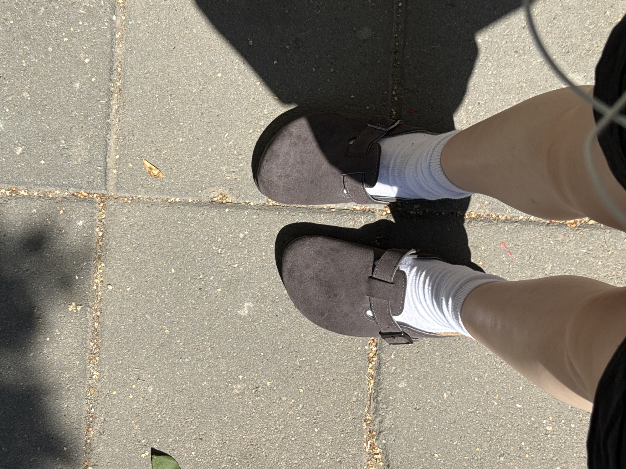 A top-down view shows a person's legs and feet, wearing dark brown suede clogs and white socks, standing on a paved surface made of square gray concrete tiles. The paving stones are separated by thin lines filled with small debris, possibly dirt and dried leaves. Shadows from an unseen object, likely foliage, fall across the pavement and the person's feet. A single green leaf rests on the pavement to the left of the frame, and a small dried leaf is visible on one of the paving stones. The scene appears to be outdoors, possibly in a park or on a sidewalk in Belgrade, Serbia, given the characteristic paving and abundant sunlight casting distinct shadows. The perspective is from the person's eye level, looking down at their own feet.