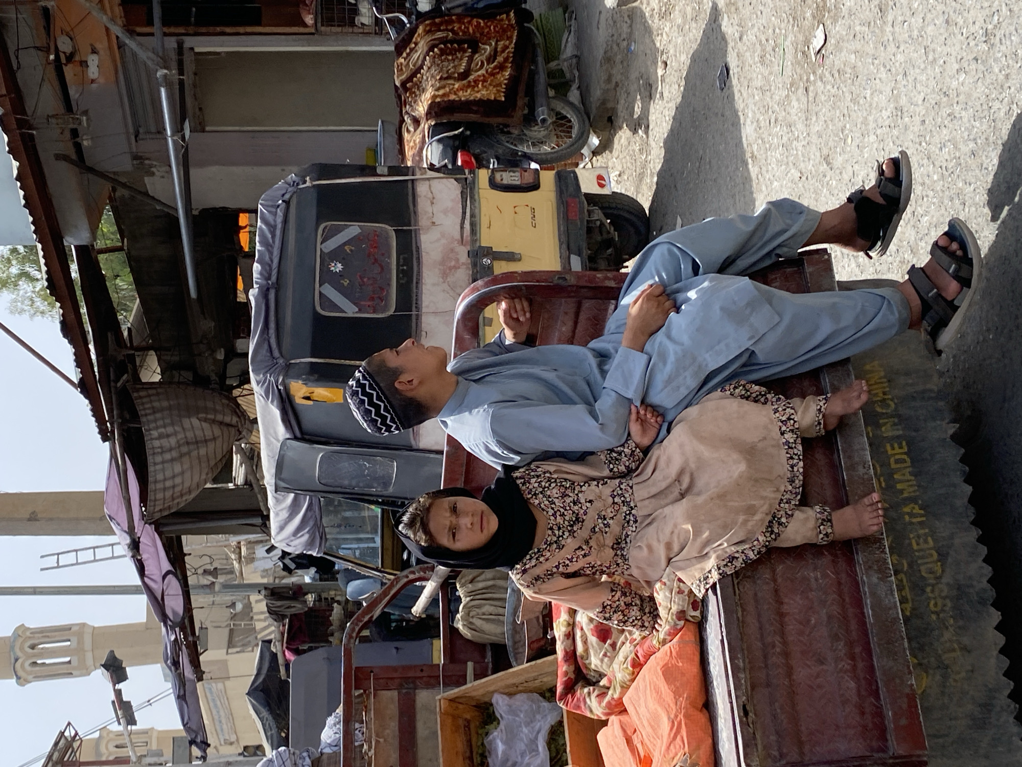 A young girl wearing a black headscarf and a light-colored, floral patterned dress sits on the edge of a red truck bed next to a boy wearing a light blue shalwar kameez and a black and white kufi. The girl is looking towards the camera, while the boy is looking to his left. They are both sitting on blankets and other items in the back of the truck. Behind them, a yellow and black auto rickshaw is parked. In the background, there are buildings, a ladder leaning against a structure, and a motorcycle covered with a blanket. The scene appears to be in a marketplace or street in Kandahar, Afghanistan, as suggested by the architecture and the presence of an auto rickshaw, a common mode of transport in the region. The truck bed has some text inscribed on its side, which appears to be "QUETTA. MADE IN CHINA."