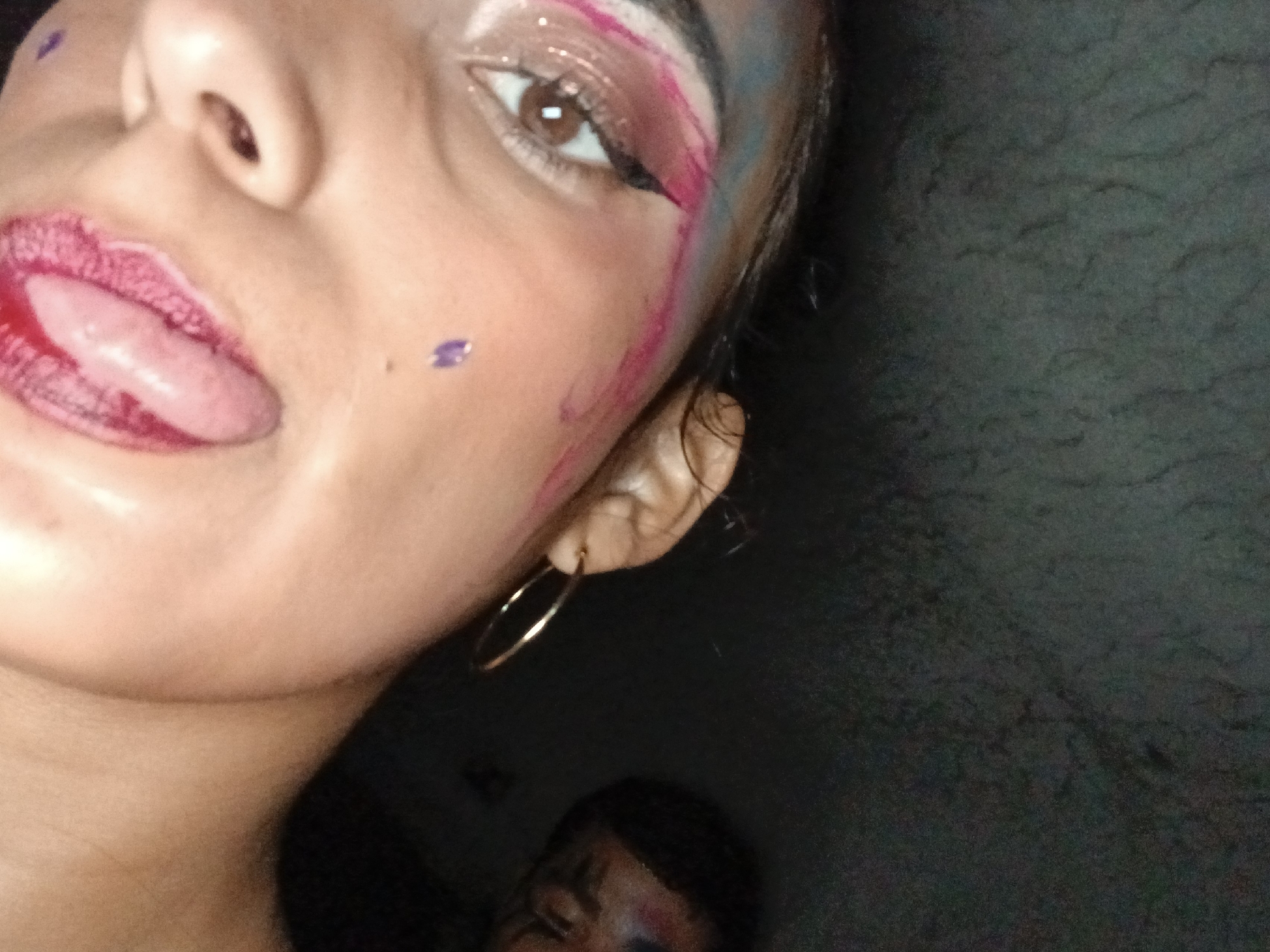 Close-up shot of a person's face with makeup, including pink and blue streaks, and a purple dot on the cheek. The person has their tongue sticking out. Gold hoop earring is visible. Another person is partially visible in the lower part of the image, also with makeup on their face. The background is dark and textured. No specific indicators link this to Playa El Agua, Venezuela.