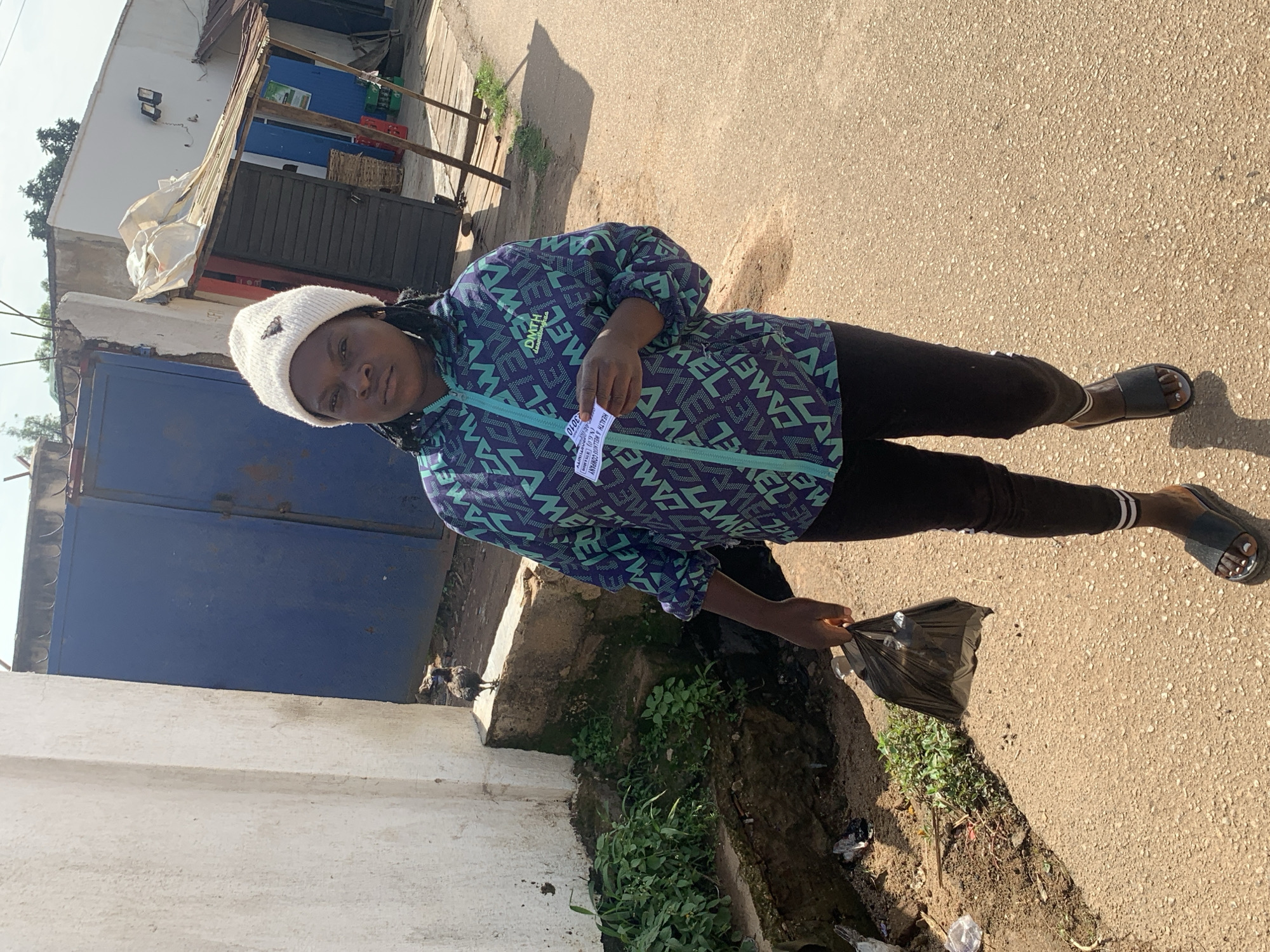 A young Nigerian woman wearing a white beanie, a colorful patterned jacket, black leggings, and black sandals walks on a dirt road. She is holding a black plastic bag in her right hand and a small white card in her left hand. A bird is perched on a concrete ledge to her left. In the background, there is a blue metal gate, a white building with a corrugated metal roof, and some vegetation. The scene appears to be in a street in Jos, Nigeria.