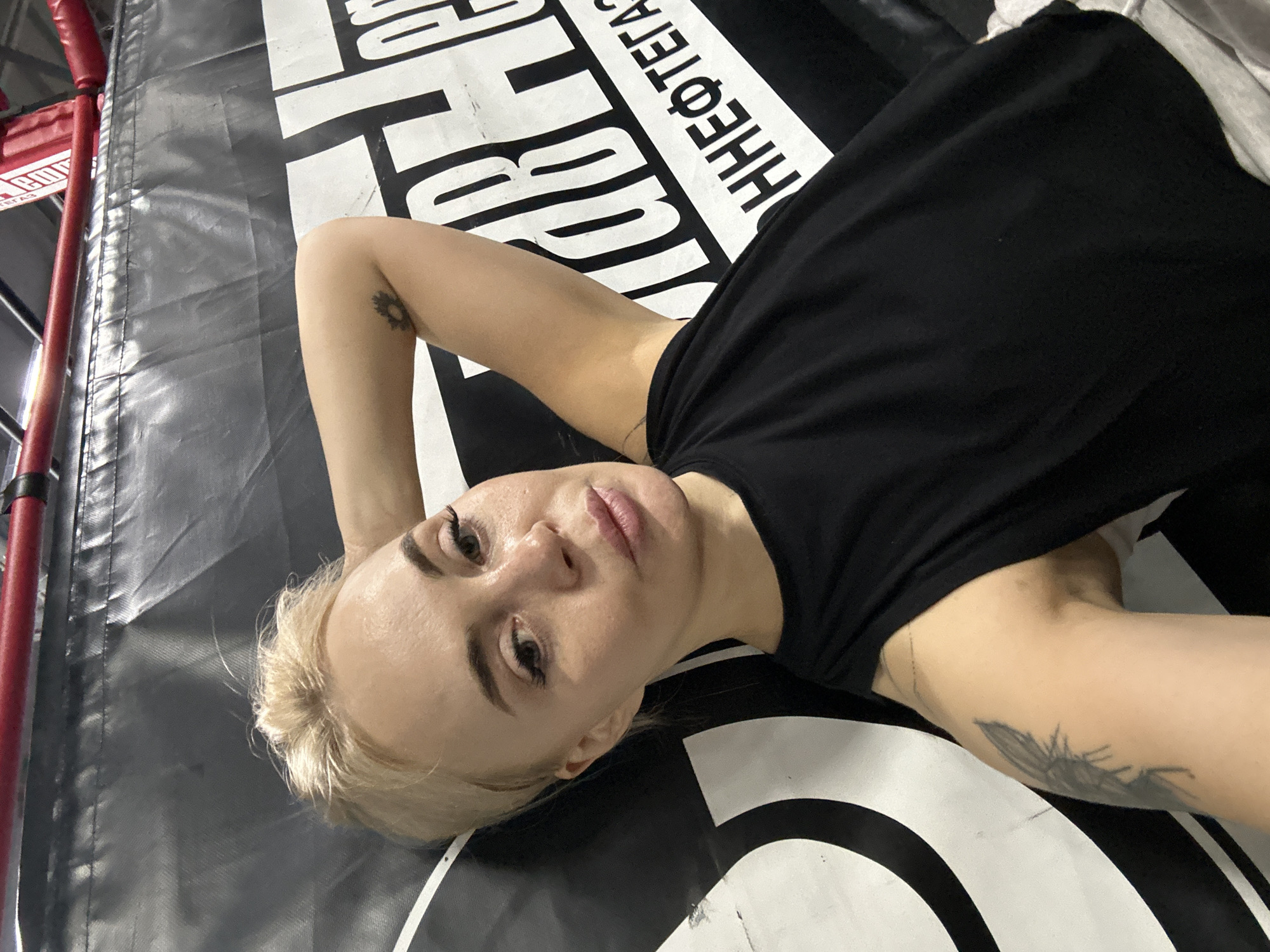 A woman with short blonde hair, wearing a black top, is lying on a surface with black and white text. She has a tattoo on her left forearm and another tattoo on her right upper arm. Part of a boxing ring with red and grey padding is visible in the upper left corner. The location is potentially in Megion, Russia, based on the Cyrillic text on the surface the woman is lying on.