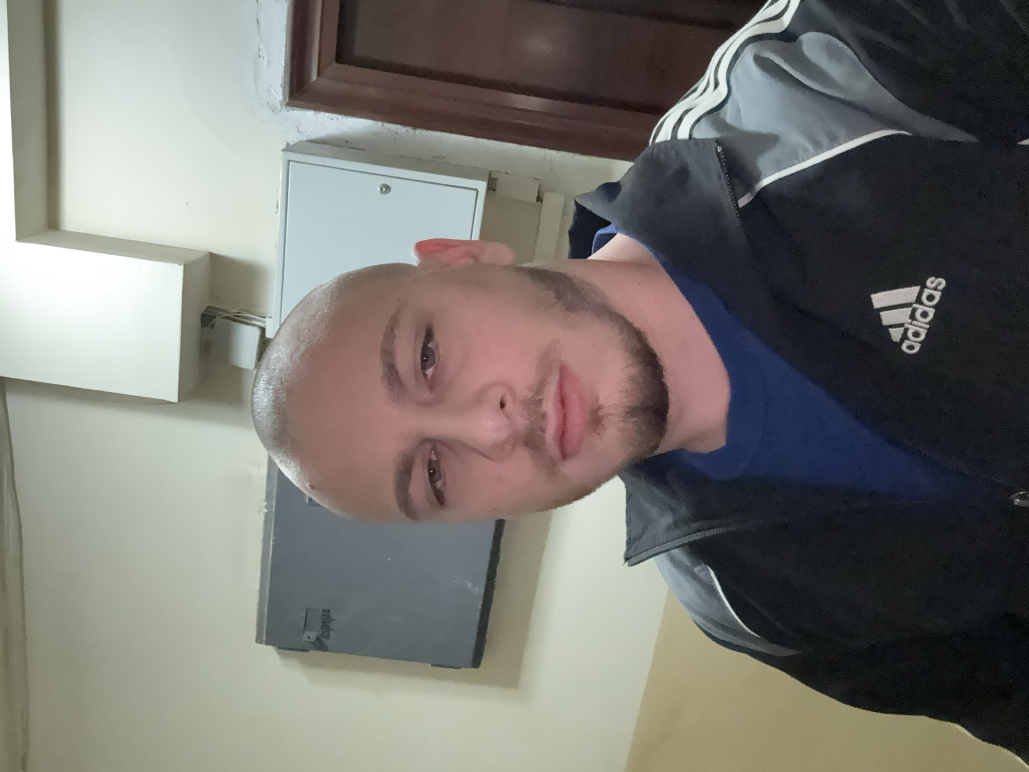 A medium shot captures a light-skinned male with a shaved head and a beard. He's wearing a blue shirt under a black Adidas jacket with grey and white accents. The setting appears to be indoors, against a wall painted in light beige. Behind the man, there's a grey electrical panel and a white box, positioned below a door frame. The image does not provide any information to allow identification of a location in the city of Stara Zagora, Bulgaria.