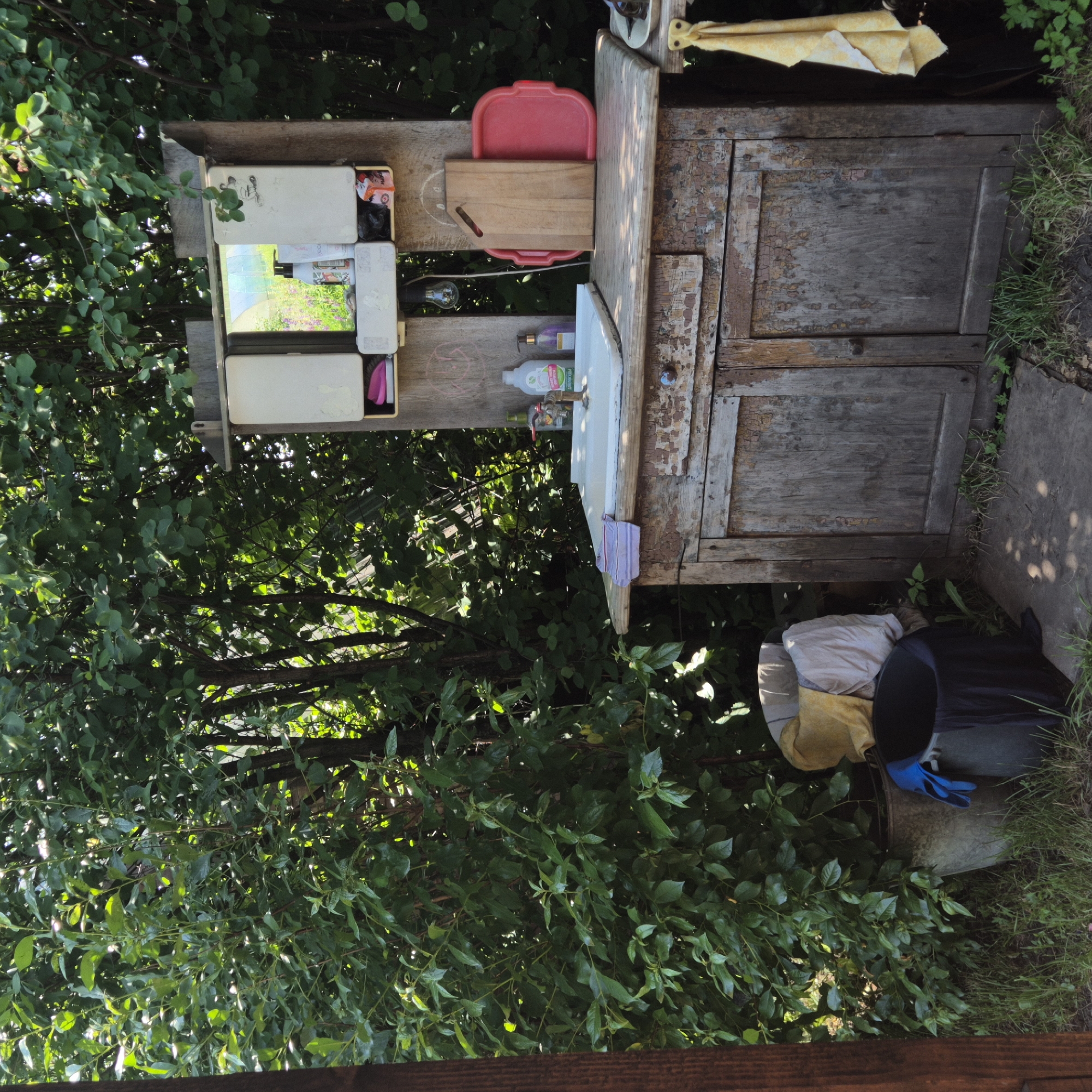 This is a medium shot, eye-level, outdoor photograph of a rustic outdoor sink setup located amidst lush green foliage in Новостройка, Russia. The sink is part of an old, weathered wooden cabinet with peeling paint, suggesting it's in a rural or garden setting. Above the sink, there's a small mirrored cabinet with its doors open, revealing various items inside, including bottles of soap or lotion, and a pink slipper. A light bulb hangs from the cabinet. On the countertop, next to the sink, are two bottles of cleaning liquid, one white and one purple, and a faucet. A wooden cutting board and a red plastic tray are positioned to the right of the sink. In front of the cabinet, two buckets are visible; one is a larger grey metal bucket containing laundry, and a smaller black plastic bucket with blue gloves and dark fabric. The ground around the sink is covered in grass and dirt, with some fallen leaves. The background is dominated by dense green bushes and trees, indicating a natural, possibly overgrown environment. Sunlight filters through the leaves, creating dappled shadows on the ground and the sink area. The scene is static, with no people present, and appears to be a functional, albeit unkempt, outdoor washing station.