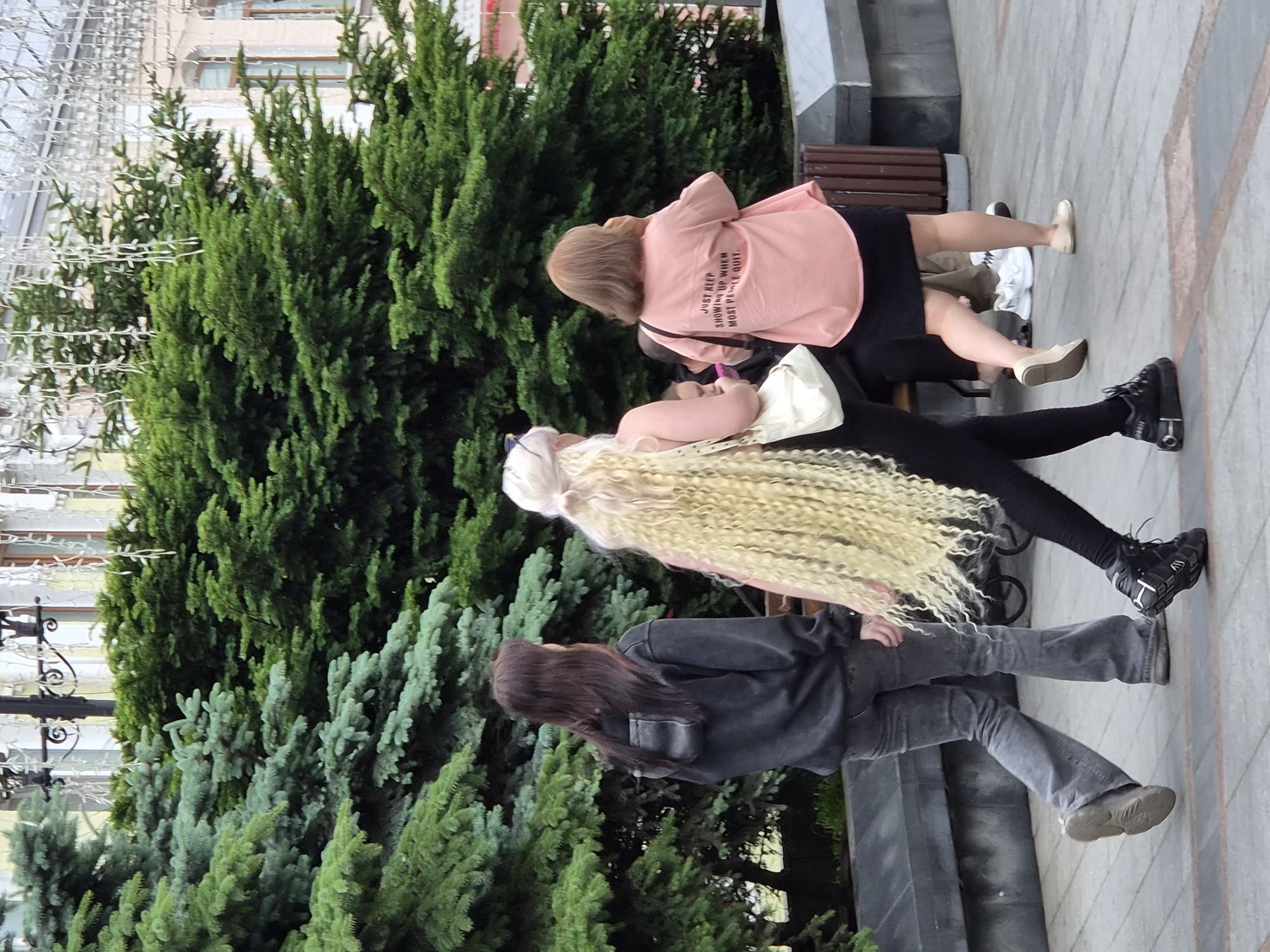 Three individuals are walking on a pedestrian walkway in Vladivostok, Russia. The first person, from left to right, is a female with long dark hair, wearing a dark grey hooded sweatshirt and grey flared jeans. She is walking to the right of the frame. The second person is a female with very long blonde curly hair, wearing a black tank top and black pants. She is walking slightly ahead of the first person, also to the right. She is carrying a white bag on her left shoulder. The third person is a female with short brown hair, wearing a light pink shirt with text on the back and a black skirt. She is walking behind and to the right of the second person. The walkway is paved with light grey and reddish-brown tiles. Behind the individuals are lush green evergreen trees, and further in the background, there are buildings with light-colored facades and windows. Decorative white string lights are visible hanging from a structure above the trees. A dark grey bench is situated to the right of the walkway, and a wooden trash receptacle is next to it. The scene appears to be outdoors in a city environment.