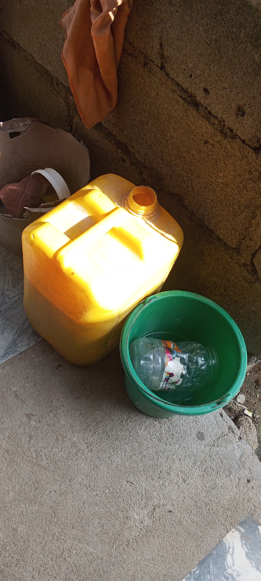 The image captures an outdoor scene in Jalingo, Nigeria. In the foreground, a rough concrete surface is visible, with a cracked pattern running across it. To the right of the concrete, a green plastic bucket sits filled with water. Inside the bucket, a clear plastic bottle with a colorful label is submerged. Behind the bucket, a large, yellow plastic container with a handle and an open spout is prominent. This container is positioned on a surface that transitions from the concrete to a tiled area. In the upper left quadrant of the image, a brown, torn cardboard container holds a piece of brown fabric with a white rim. Finally, a piece of orange cloth is draped over a textured, brown, cracked wall in the background. The scene is illuminated by sunlight, casting shadows on the various objects.